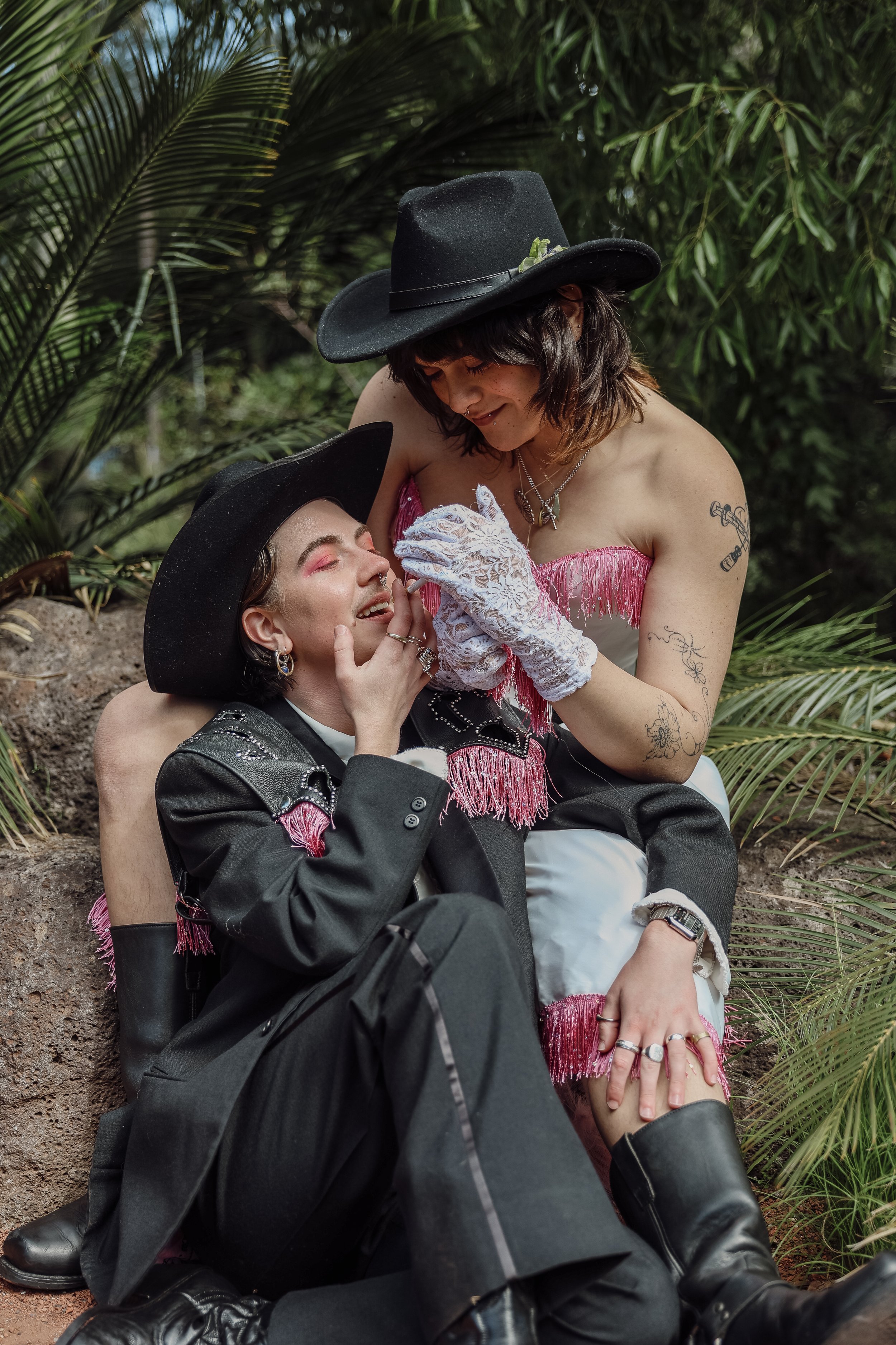 Two women dressed in flamboyant western-style outfits, with black hats and pink fringe details, are in an outdoor lush green setting, sharing an intimate moment.