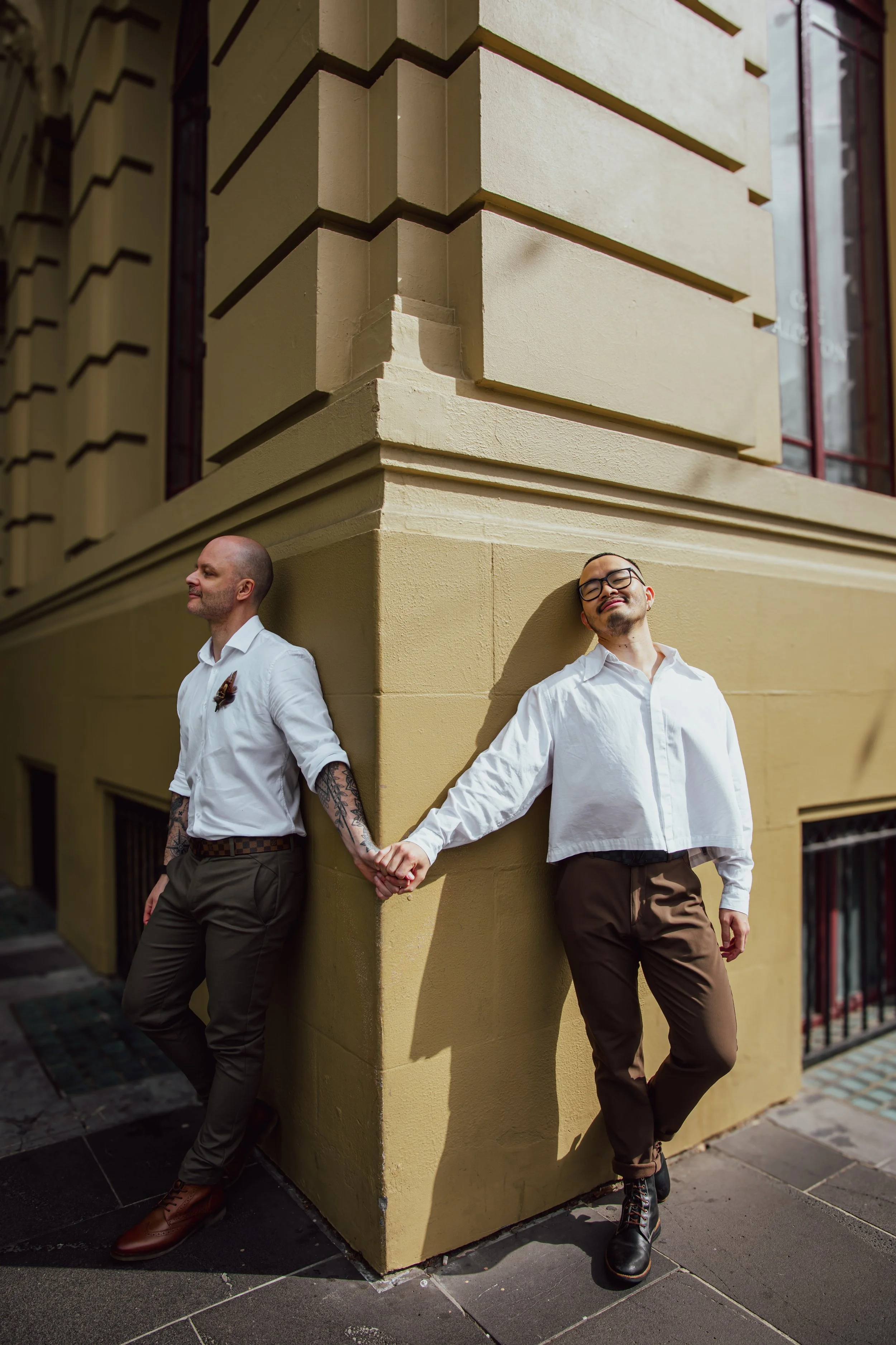 Two men holding hands while leaning against a corner of a yellow building, with one man smiling and the other with a neutral expression.