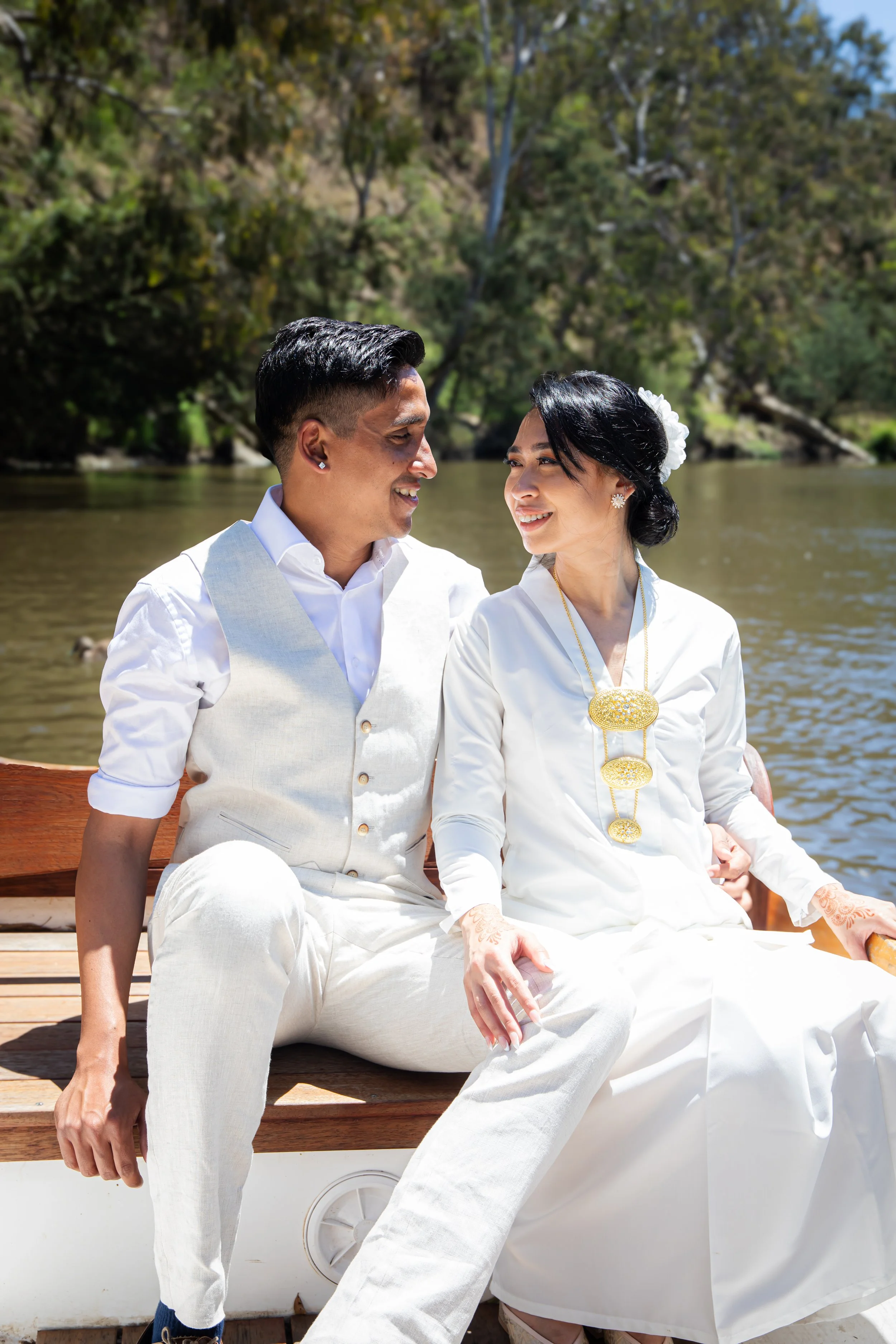 A couple in white traditional attire sitting together on a boat, smiling at each other near a river with trees in the background.