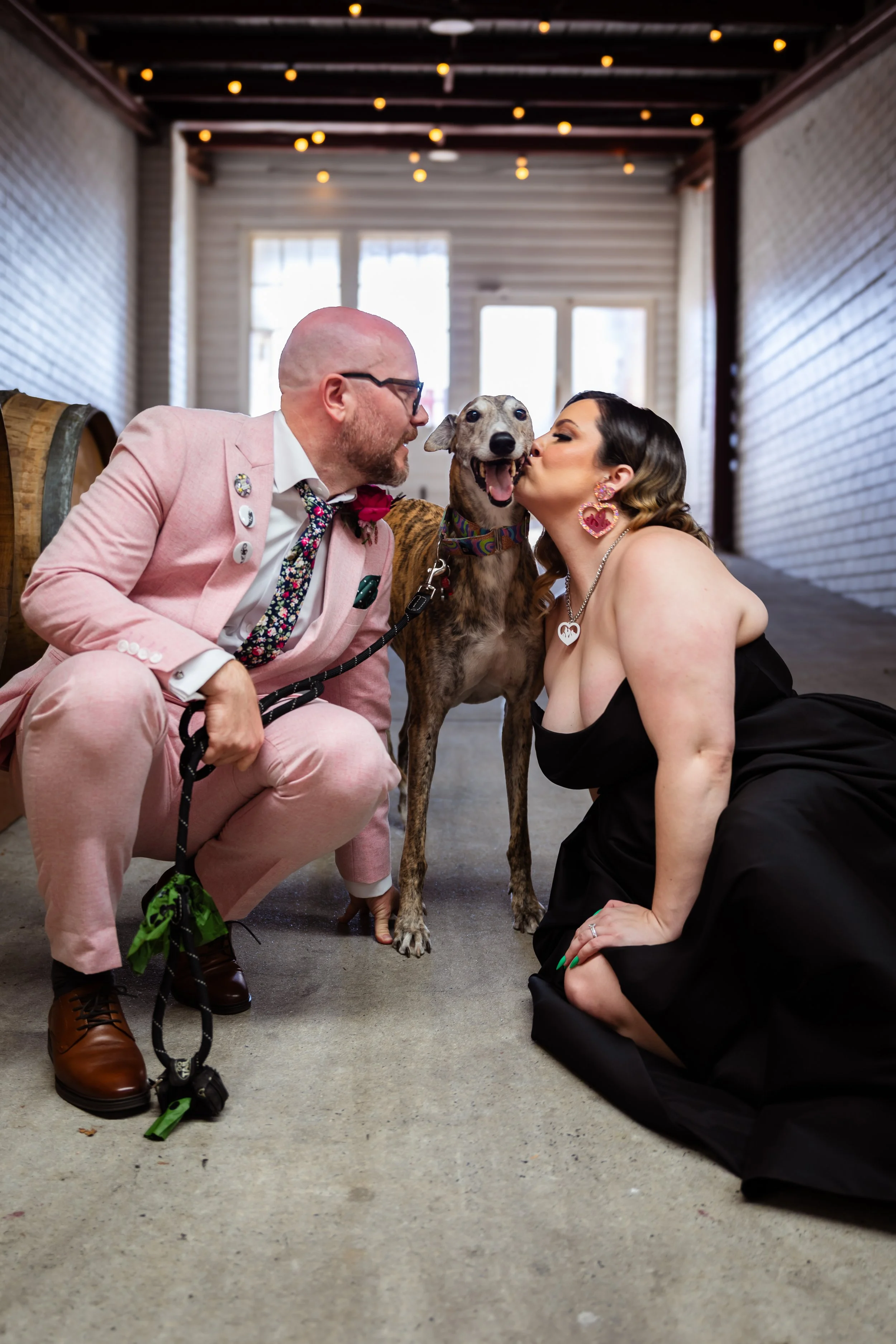 A man and woman kneeling on the floor, kissing a happy dog in the middle, in a rustic indoor setting with string lights overhead.