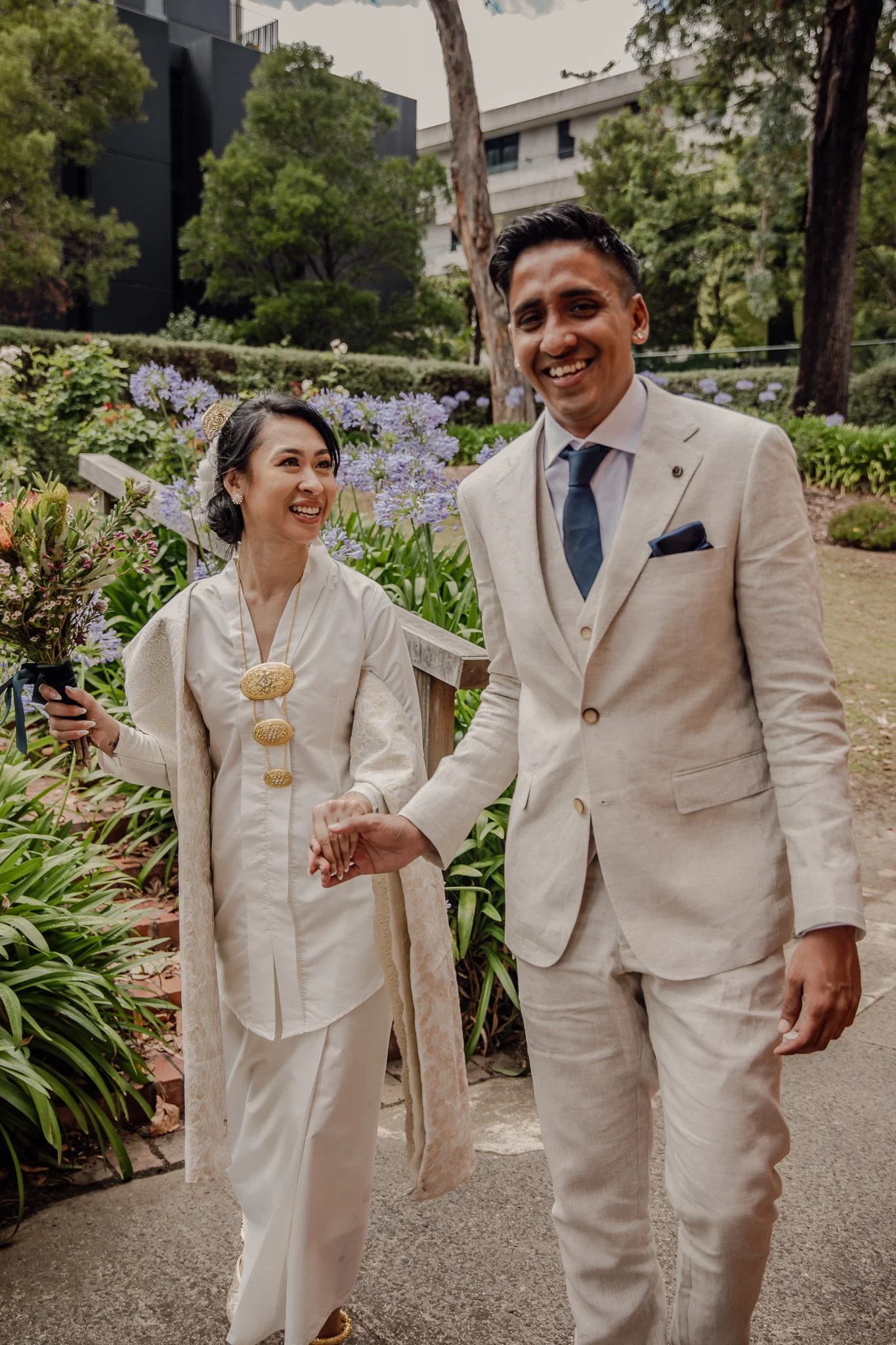A smiling couple dressed in formal attire holding hands in a garden with purple flowers and trees.