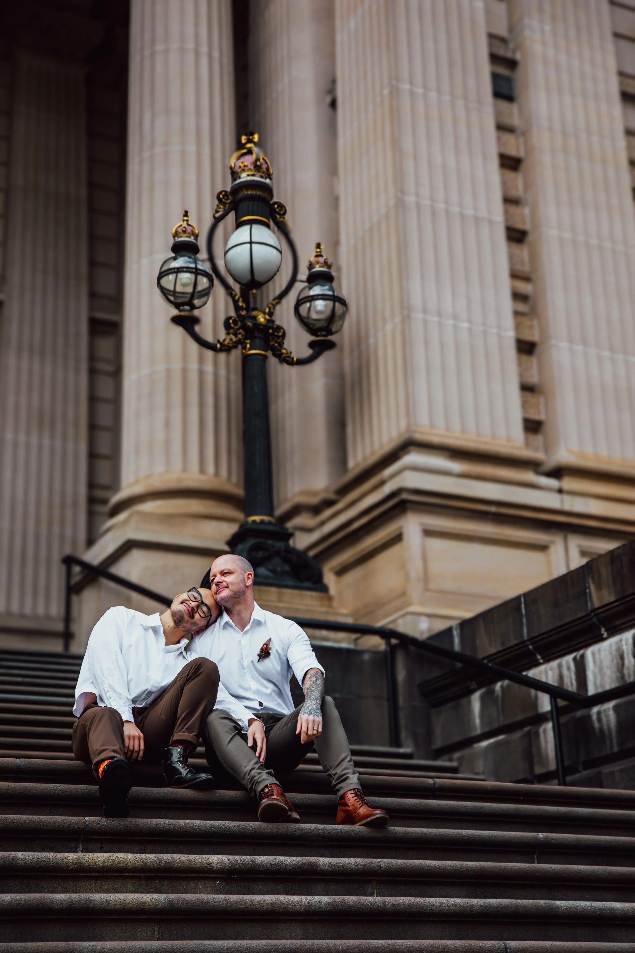 Two men sitting on stairs in front of a large historic building with a statue and a decorative lamp post, one leaning on the other's shoulder and smiling.