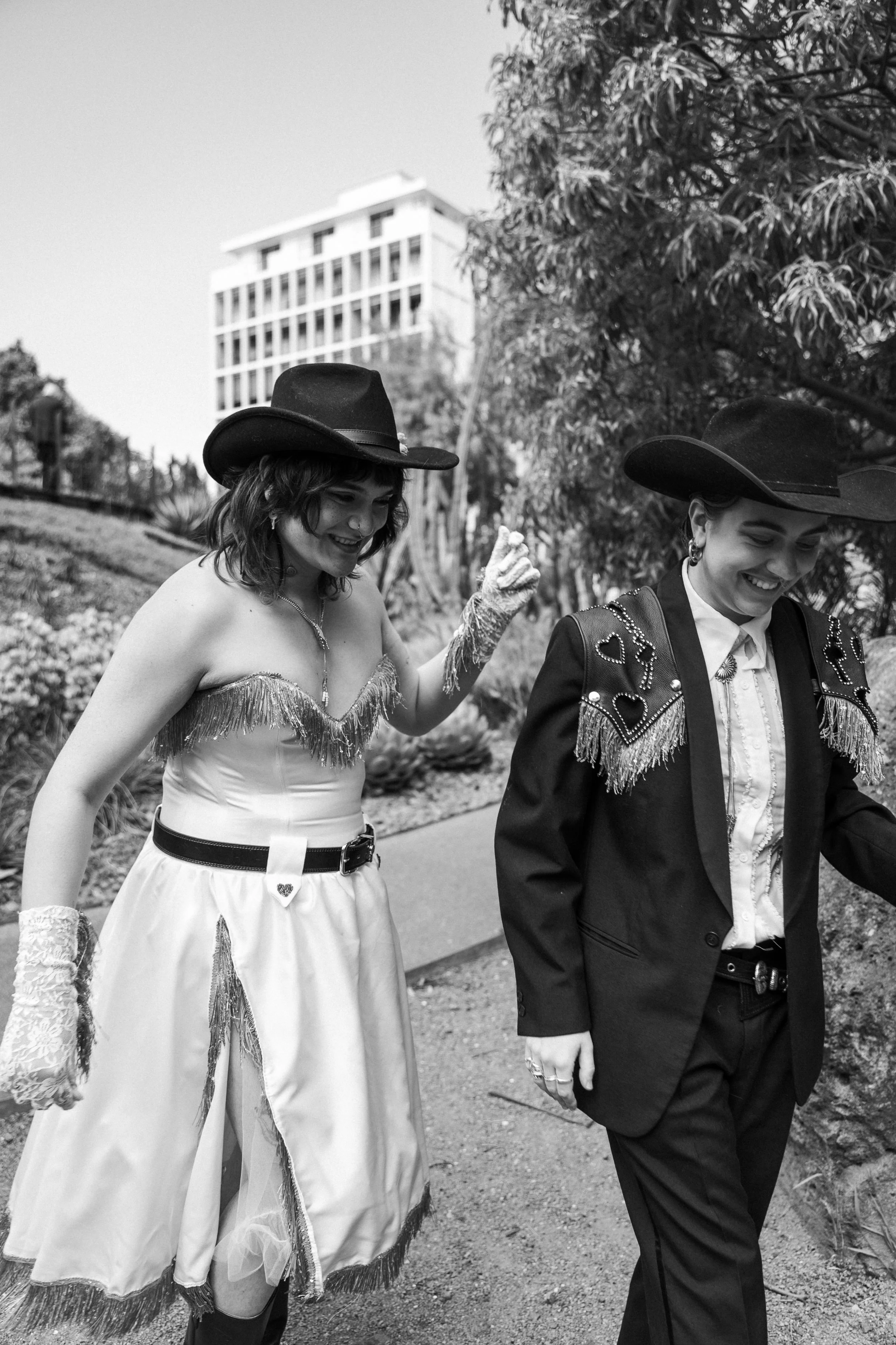 Two women dressed in cowboy-inspired outfits walking outdoors, smiling, and wearing cowboy hats.