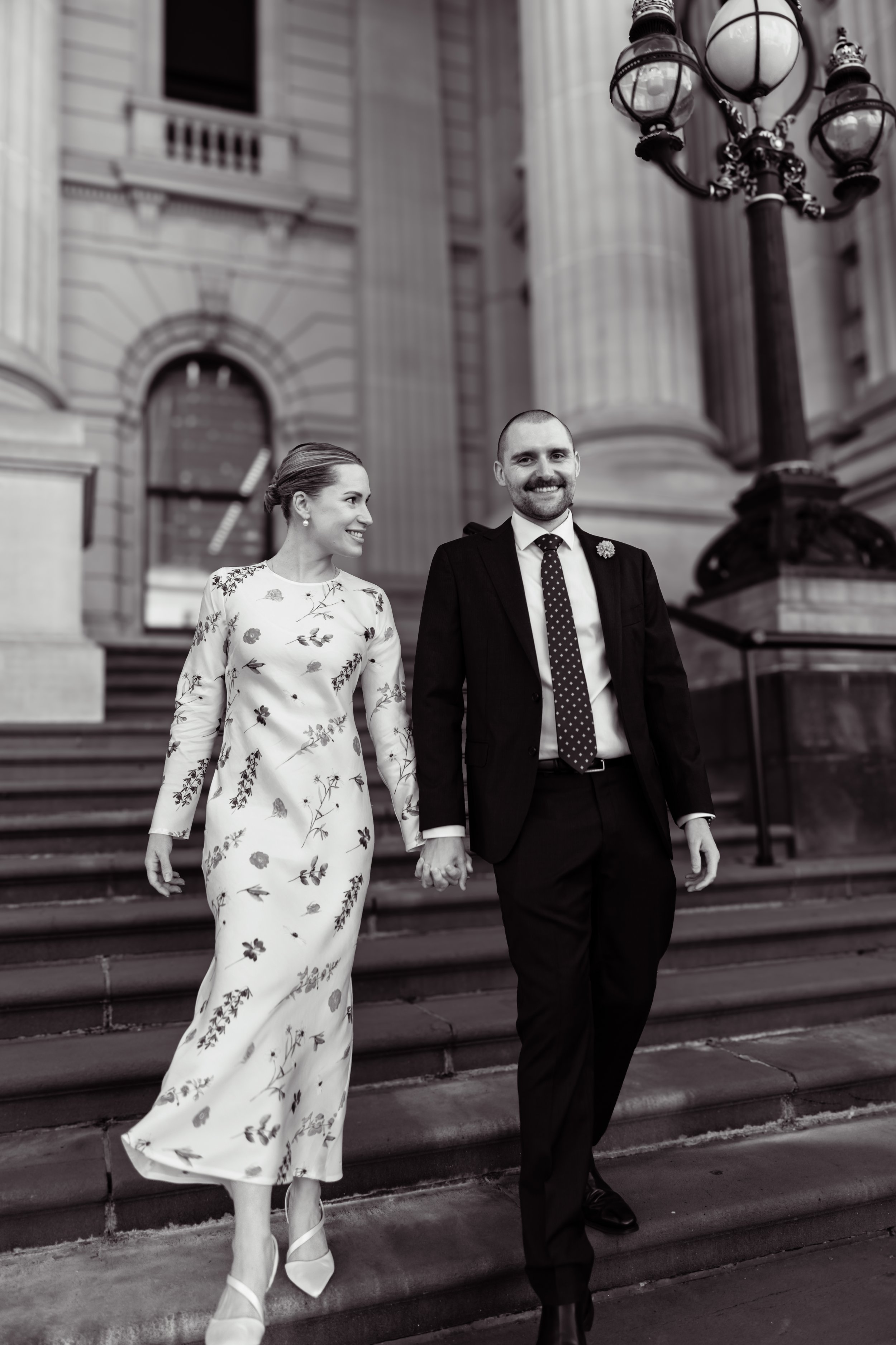 A couple holding hands, walking down outdoor stairs in front of a large historic building with columns and a street lamp, dressed in formal attire.