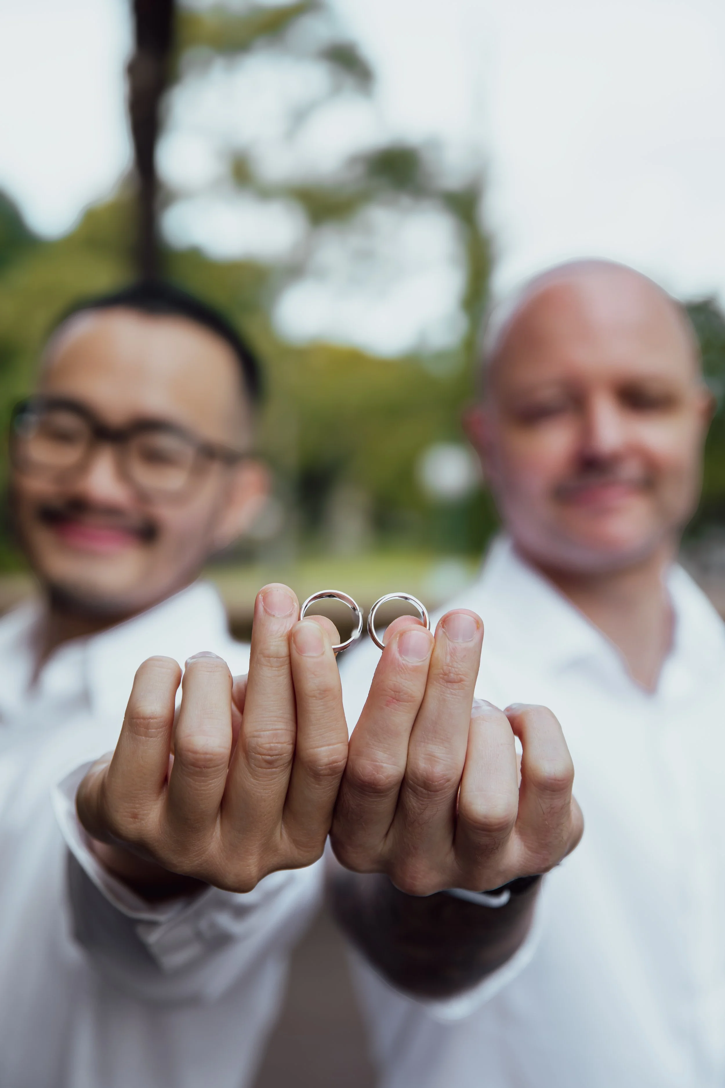 Two men holding wedding rings outdoors, blurred background.