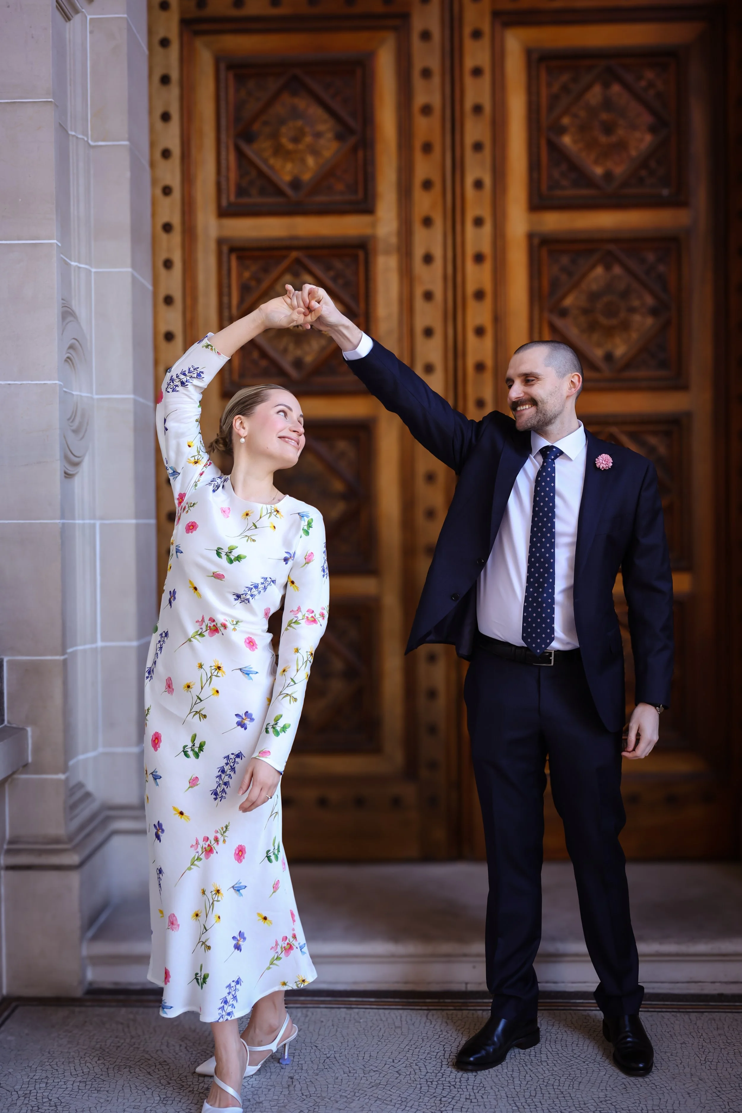 A couple dancing inside a church, holding hands up as they gaze at each other, with the woman wearing a floral dress and the man in a dark suit with a dotted tie.