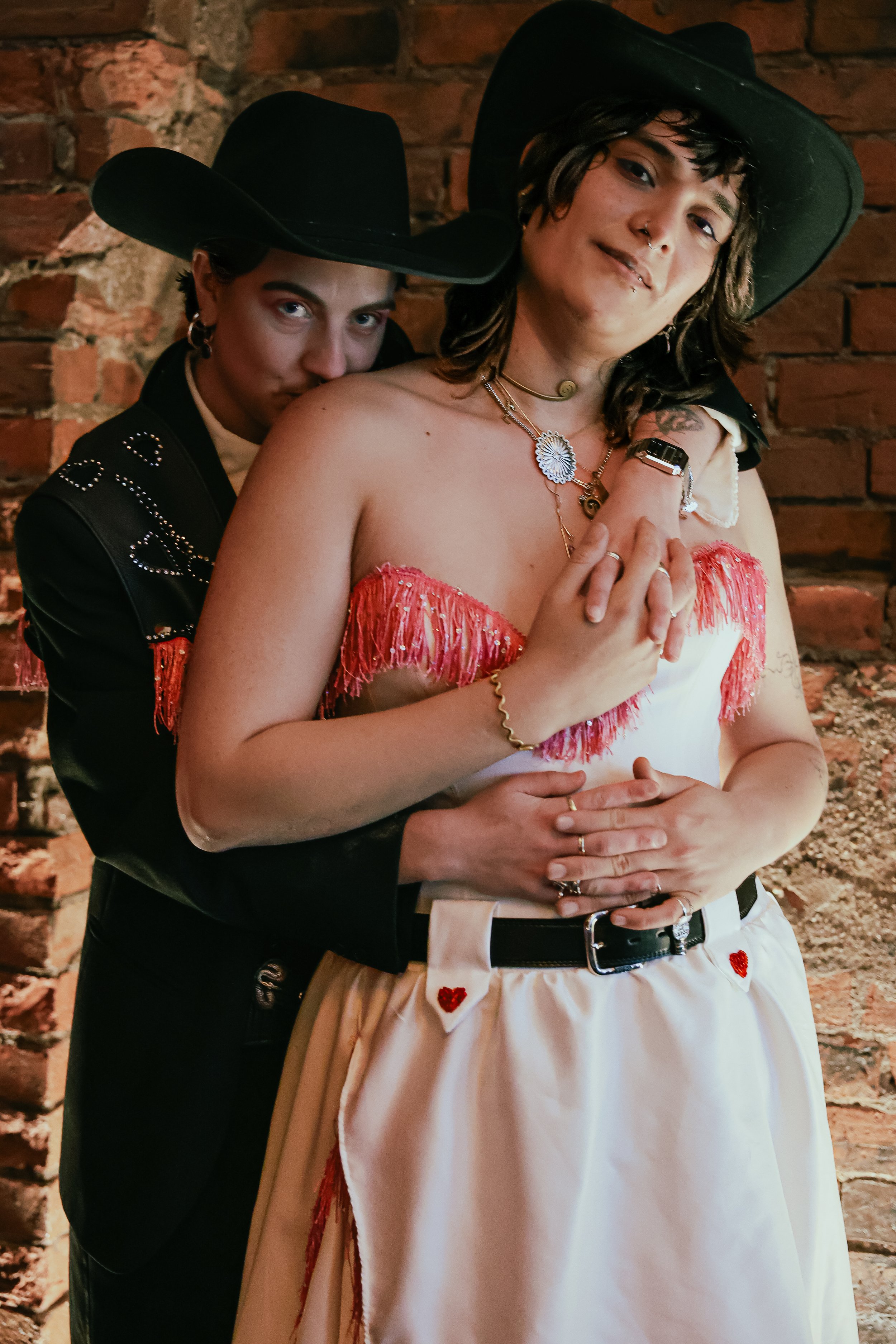 Two women wearing cowboy hats and western-style clothing, one in front and the other behind, standing against a brick wall.