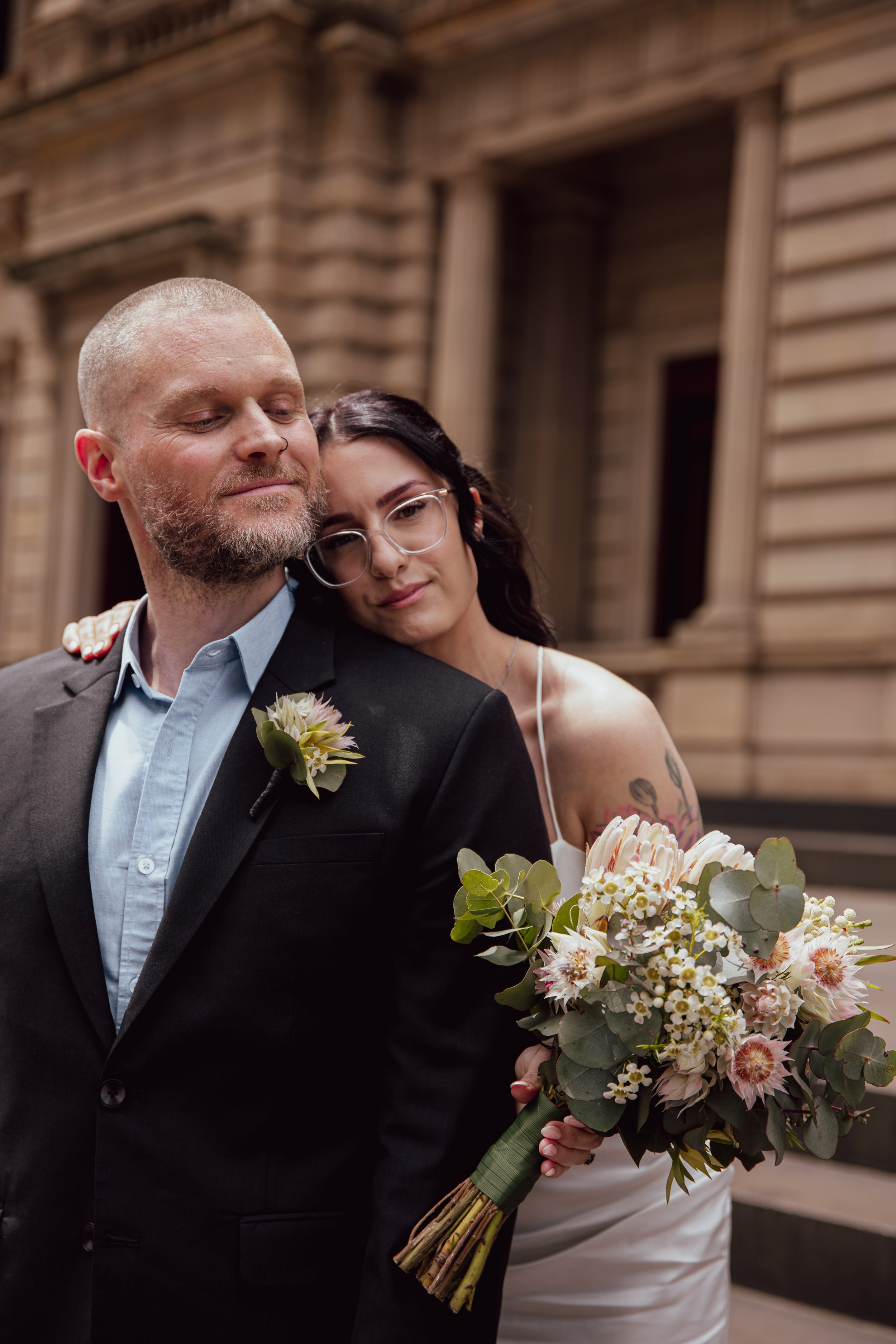A bride and groom standing close together outdoors, with the bride resting her head on the groom's shoulder, holding a bouquet of flowers. The groom is wearing a black suit with a boutonniere, and the bride is wearing a white dress with glasses, in f