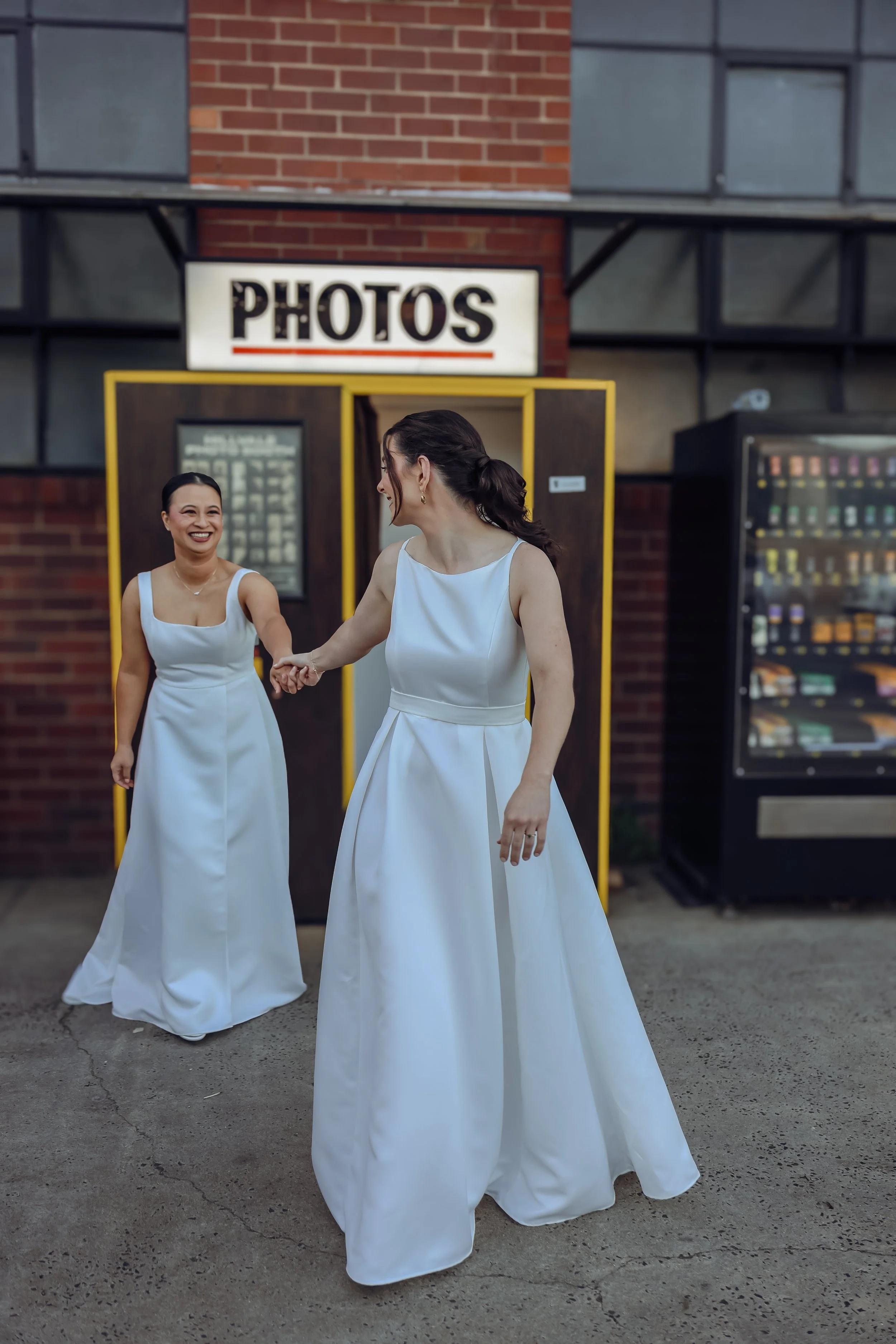 Two women in white dresses holding hands and smiling outside near a photo booth and vending machine, with a sign that says 'PHOTOS' above them.