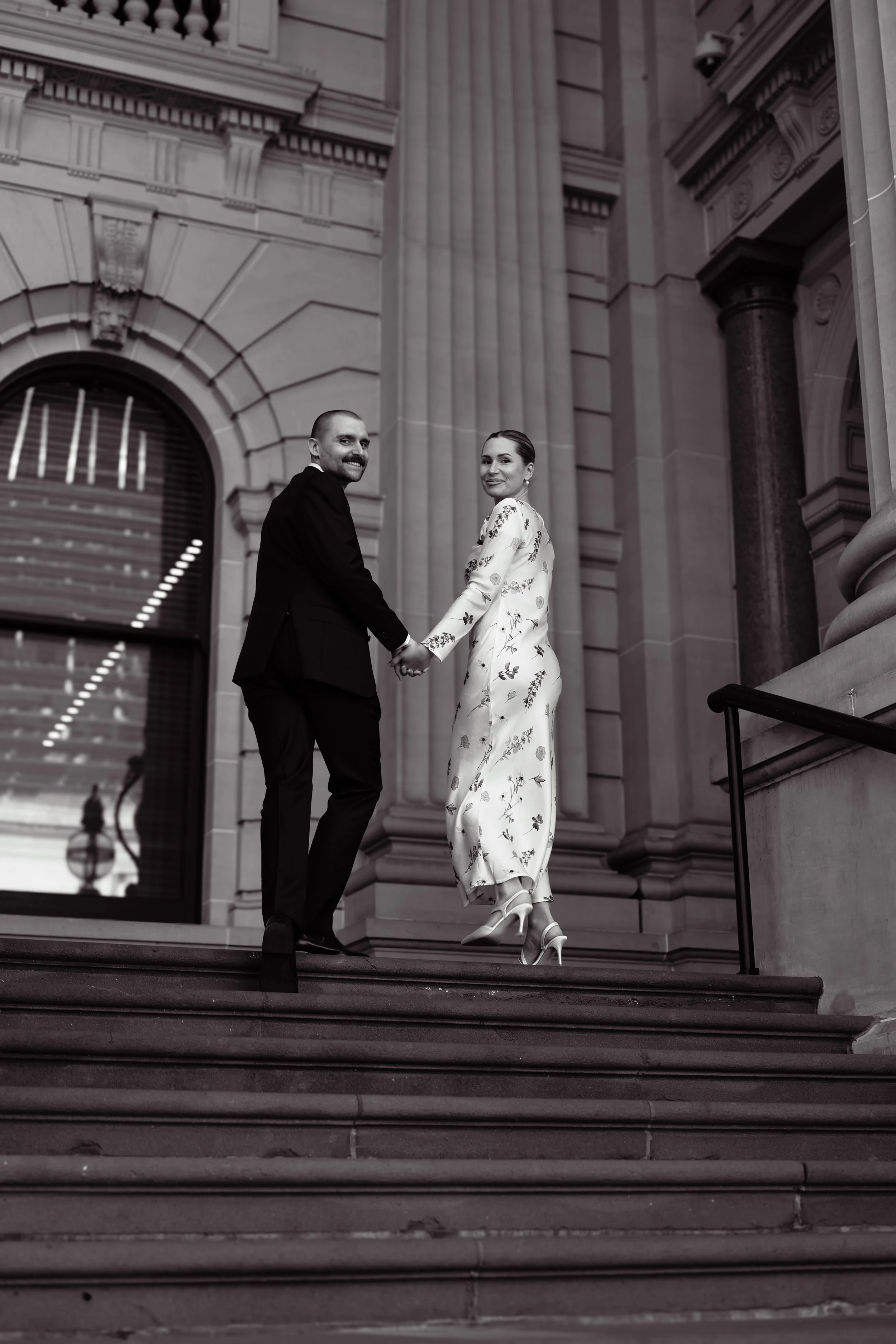 A couple in wedding attire holding hands and walking down the steps outside a grand, classical building with large columns and ornate stonework, captured in black and white.