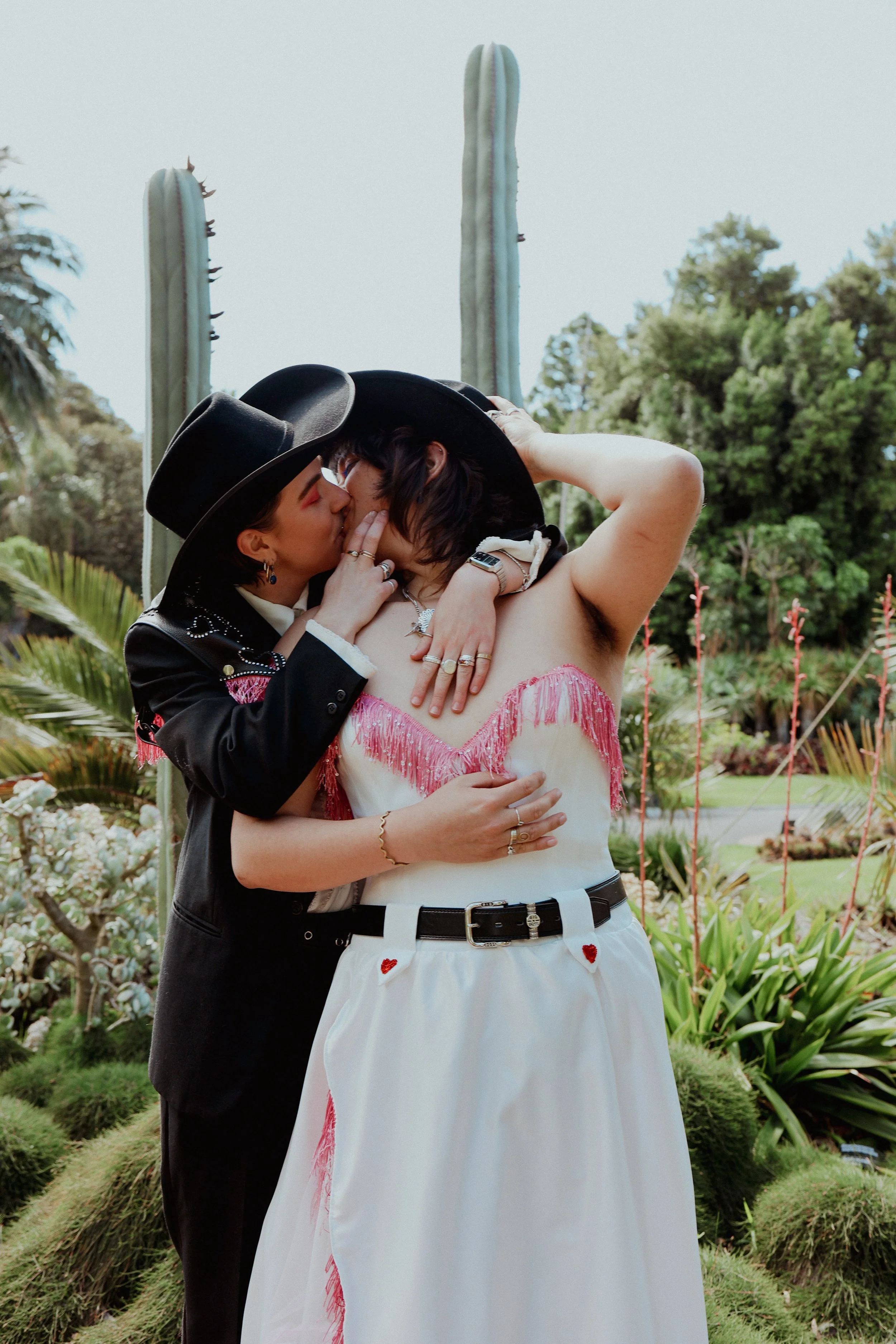 Two women sharing a kiss outdoors, one wearing a black hat and jacket, the other in a white dress with pink fringe and a black belt, surrounded by greenery and desert plants.