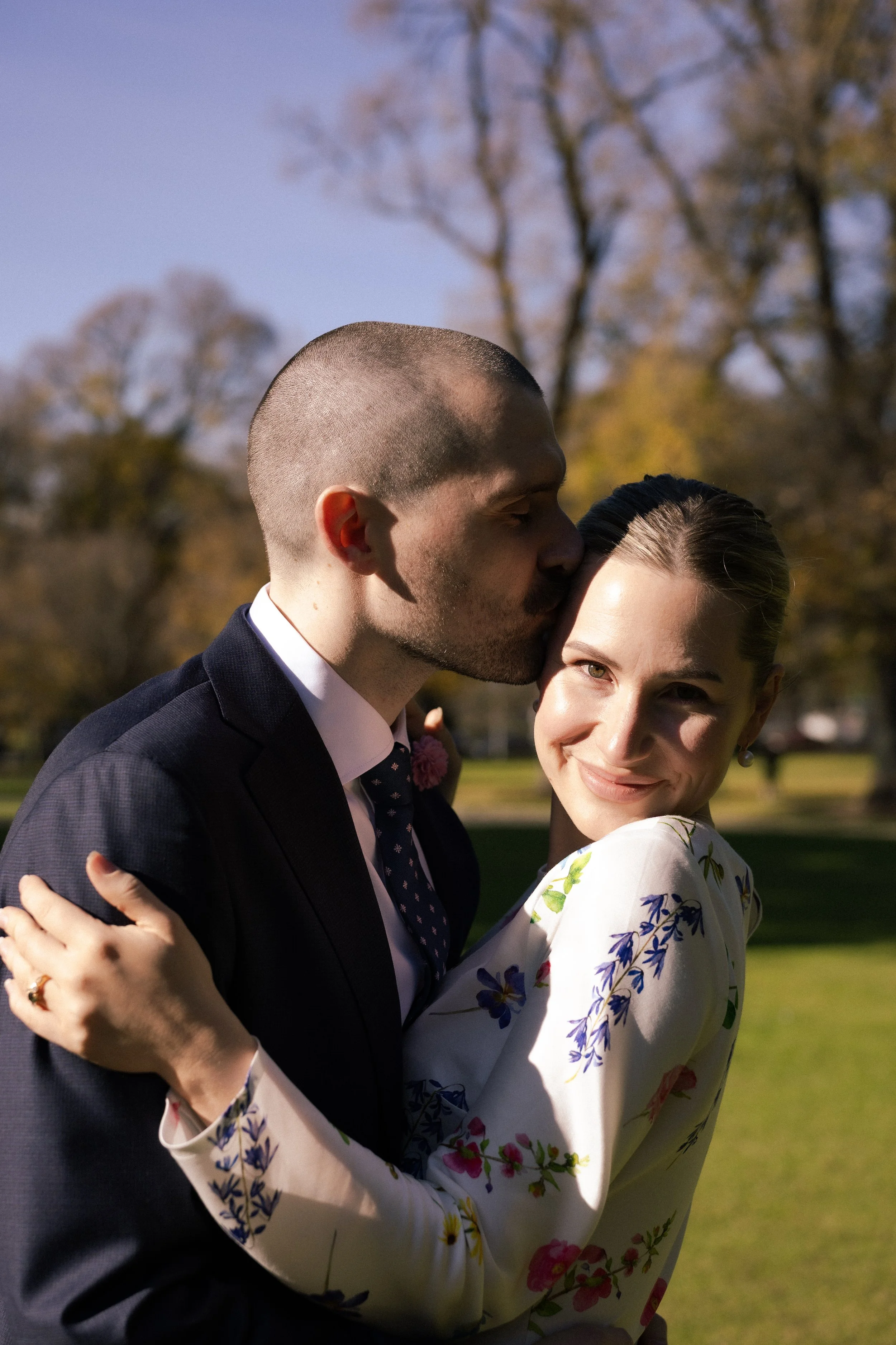 A man and woman embracing outdoors, the man kissing the woman's forehead, both smiling, with trees and grass in the background.