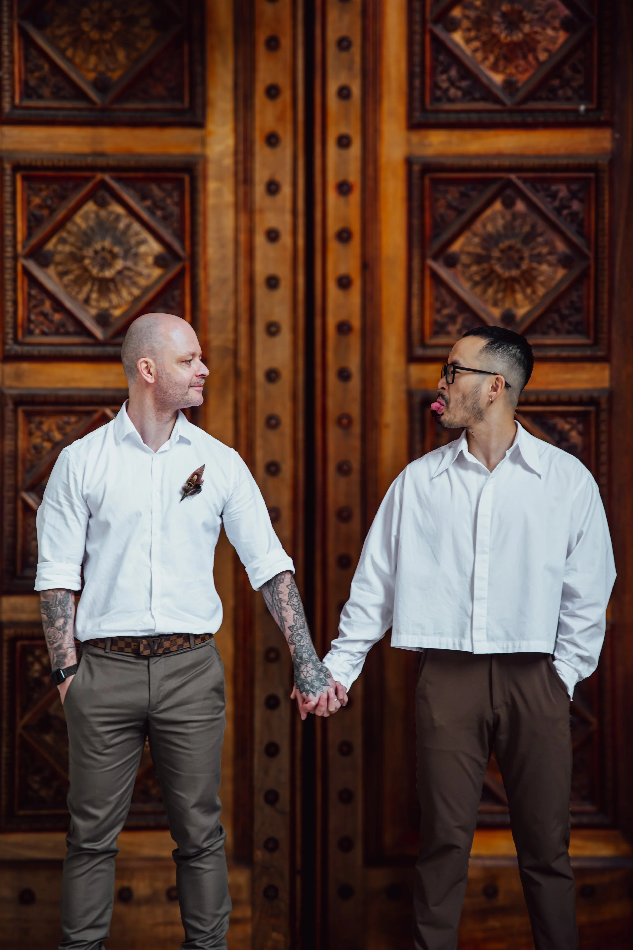 Two men holding hands in front of a large wooden door. One man has tattoos and is wearing a white shirt with a butterfly pin. The other man is wearing glasses and has a short haircut, also in a white shirt. They are making face expressions at each ot
