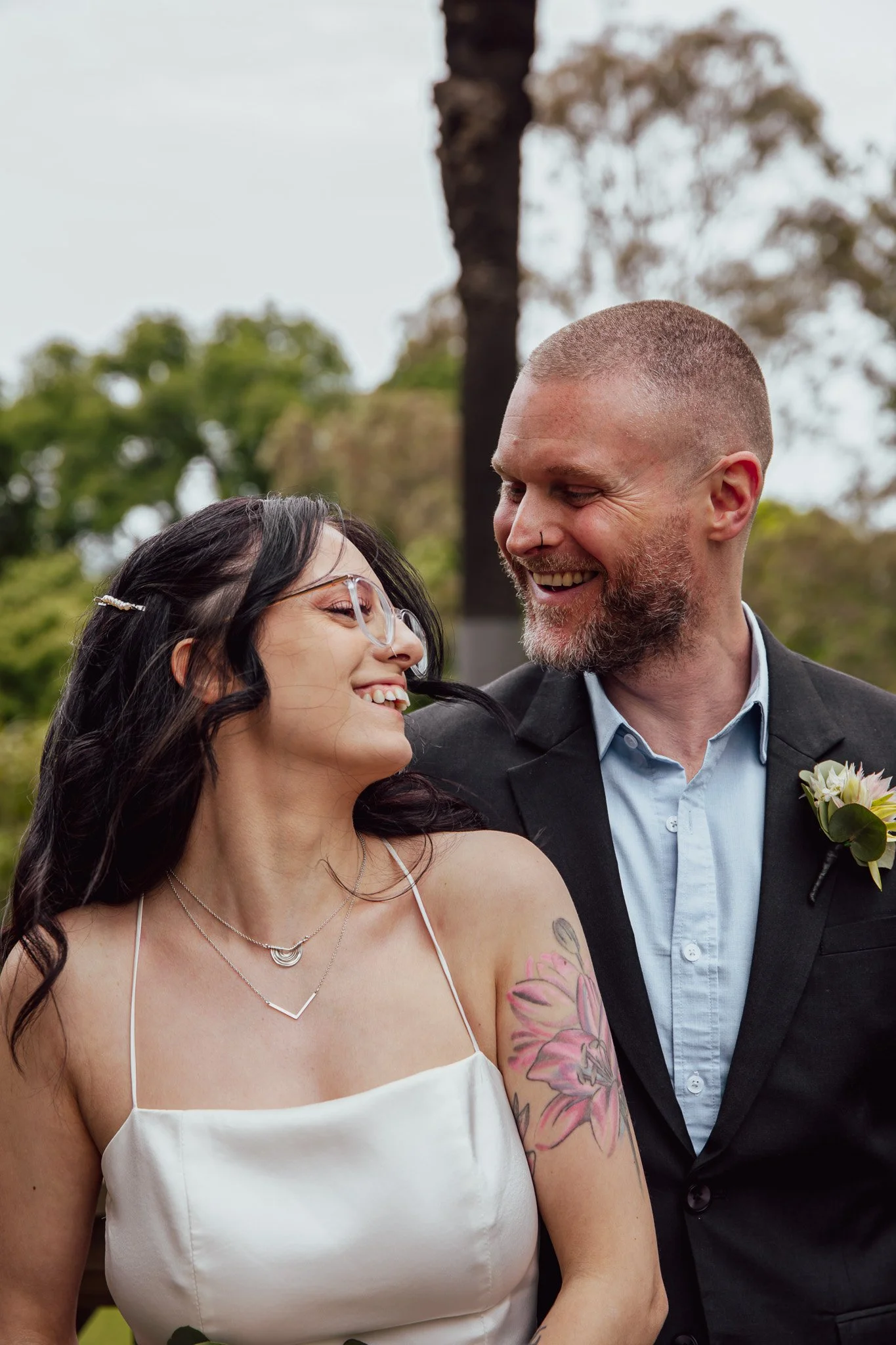 A happy couple, a woman with black hair and glasses, and a man with a beard, are smiling at each other outdoors, dressed formally for a wedding or special occasion.