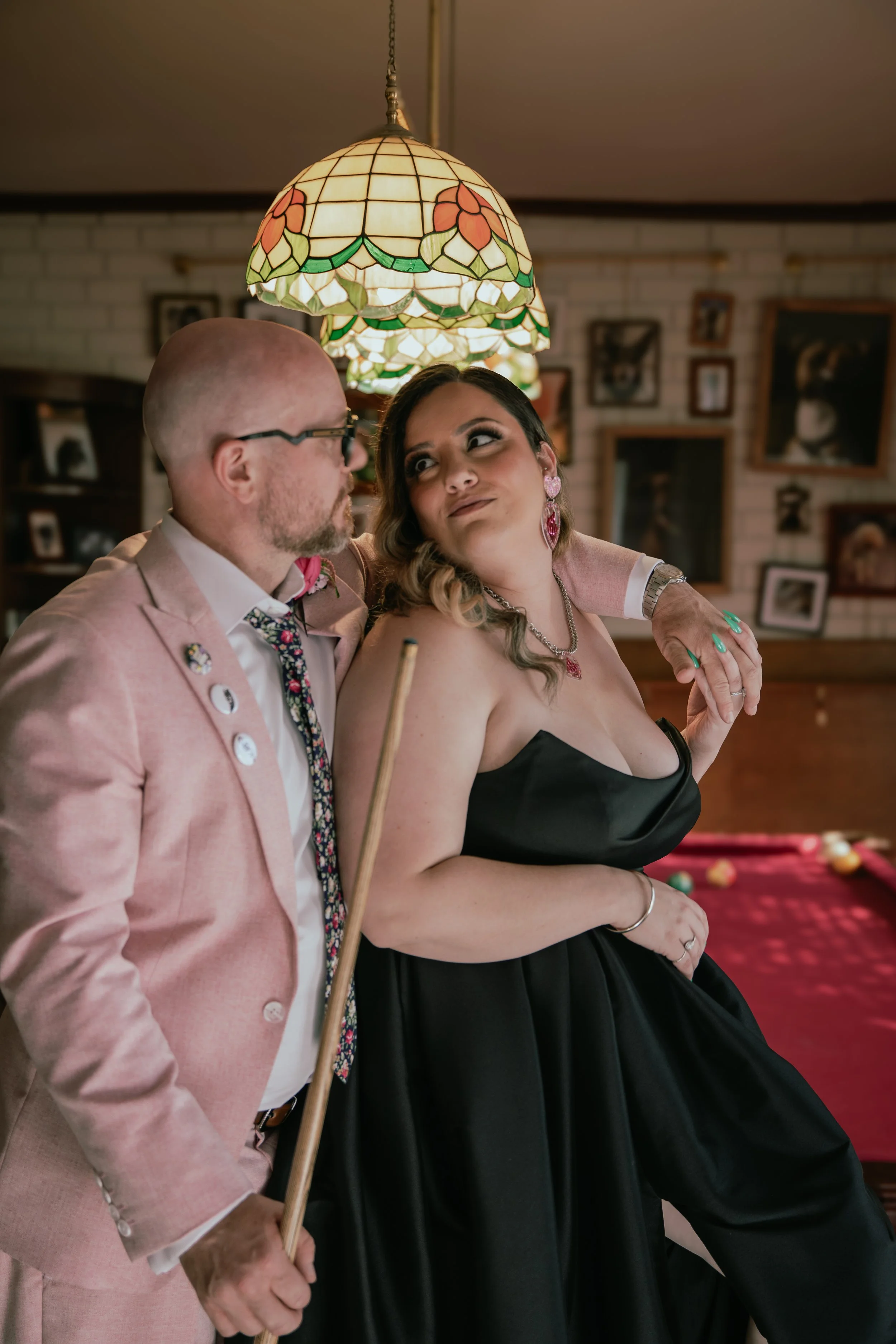 A man and woman socialize near a pool table in a cozy, vintage-themed room. The man, dressed in a pink blazer and patterned tie, holds a pool cue, while the woman, wearing a strapless black dress and colorful jewelry, leans into him, smiling.