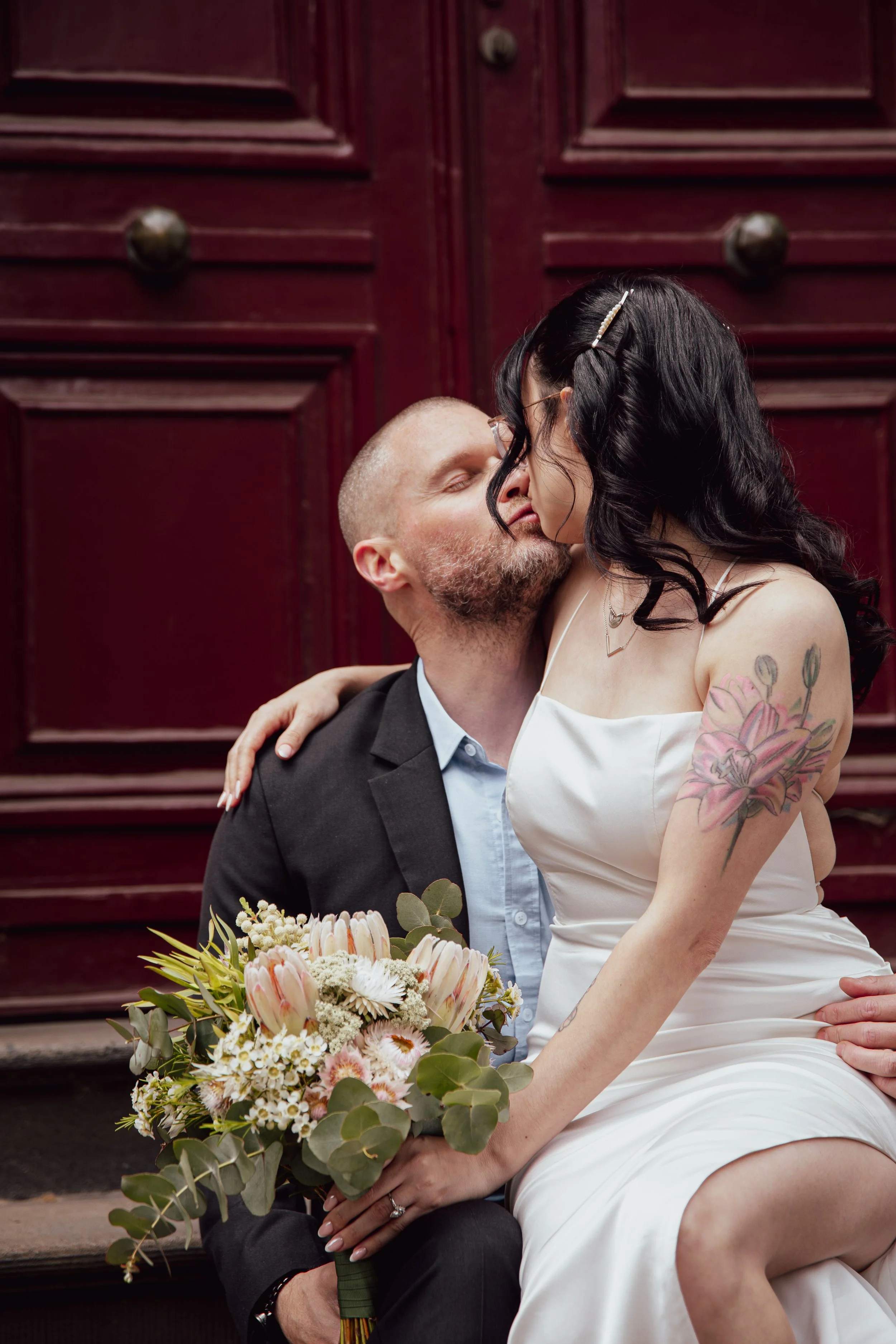A couple kissing, the woman with dark hair and a tattooed shoulder, wearing a white dress, holding a bouquet of flowers, with a man in a suit, against a red wooden door background.