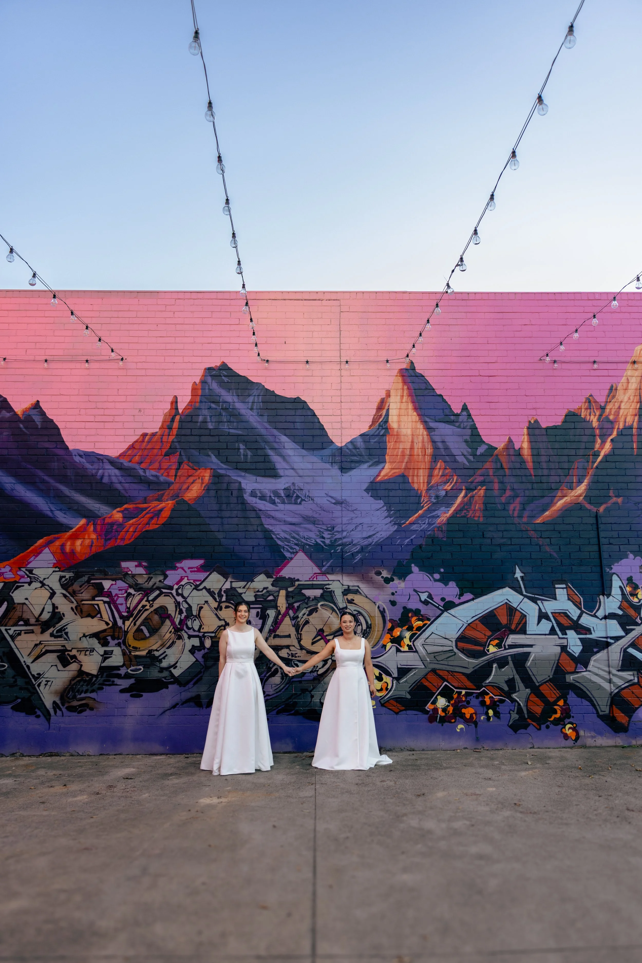Two women in white dresses holding hands in front of a colorful mountain and graffiti mural on a brick wall, with string lights overhead.