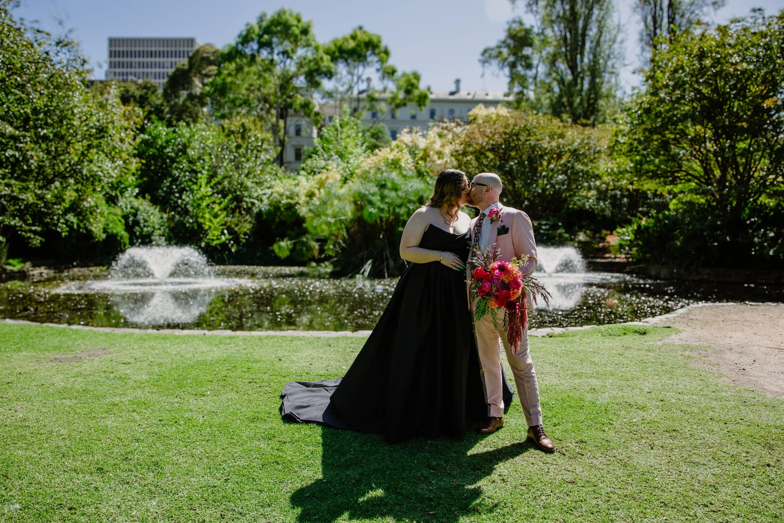 A couple in wedding attire sharing a kiss near a pond with fountains, surrounded by trees and bushes, in an urban park setting.
