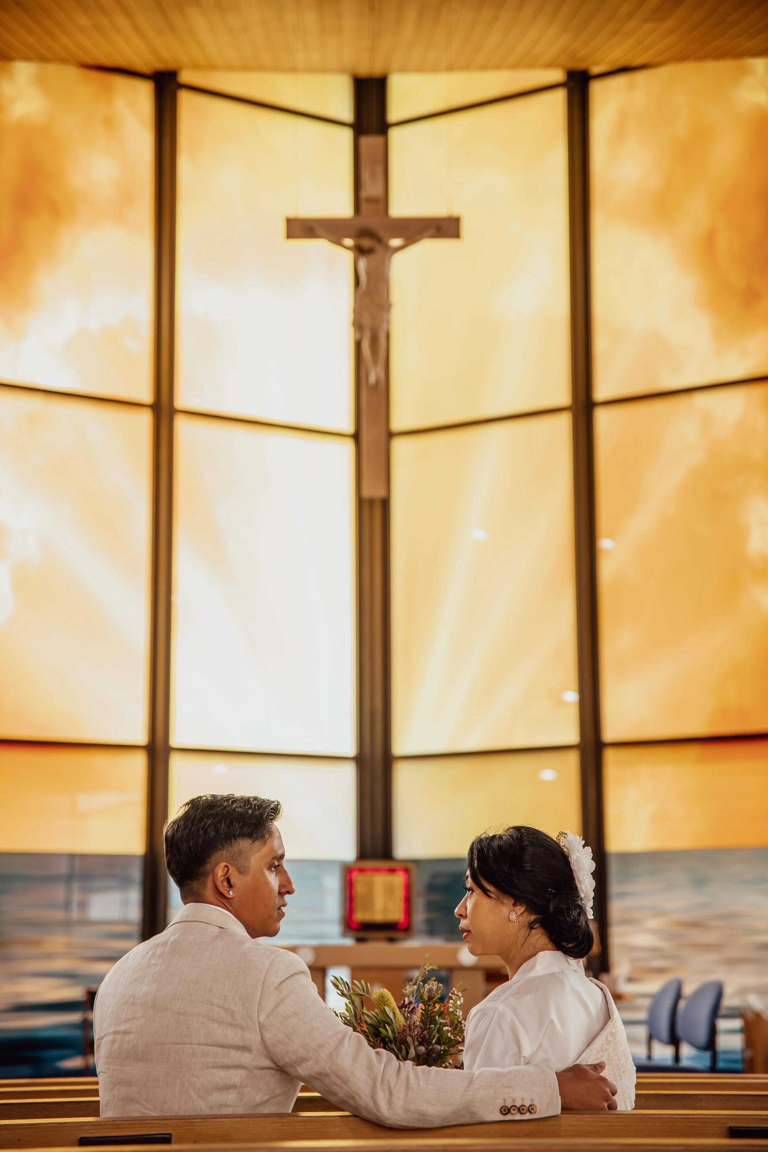 A couple sitting in a church pew during a wedding or religious ceremony, with a large crucifix and illuminated golden background behind them.