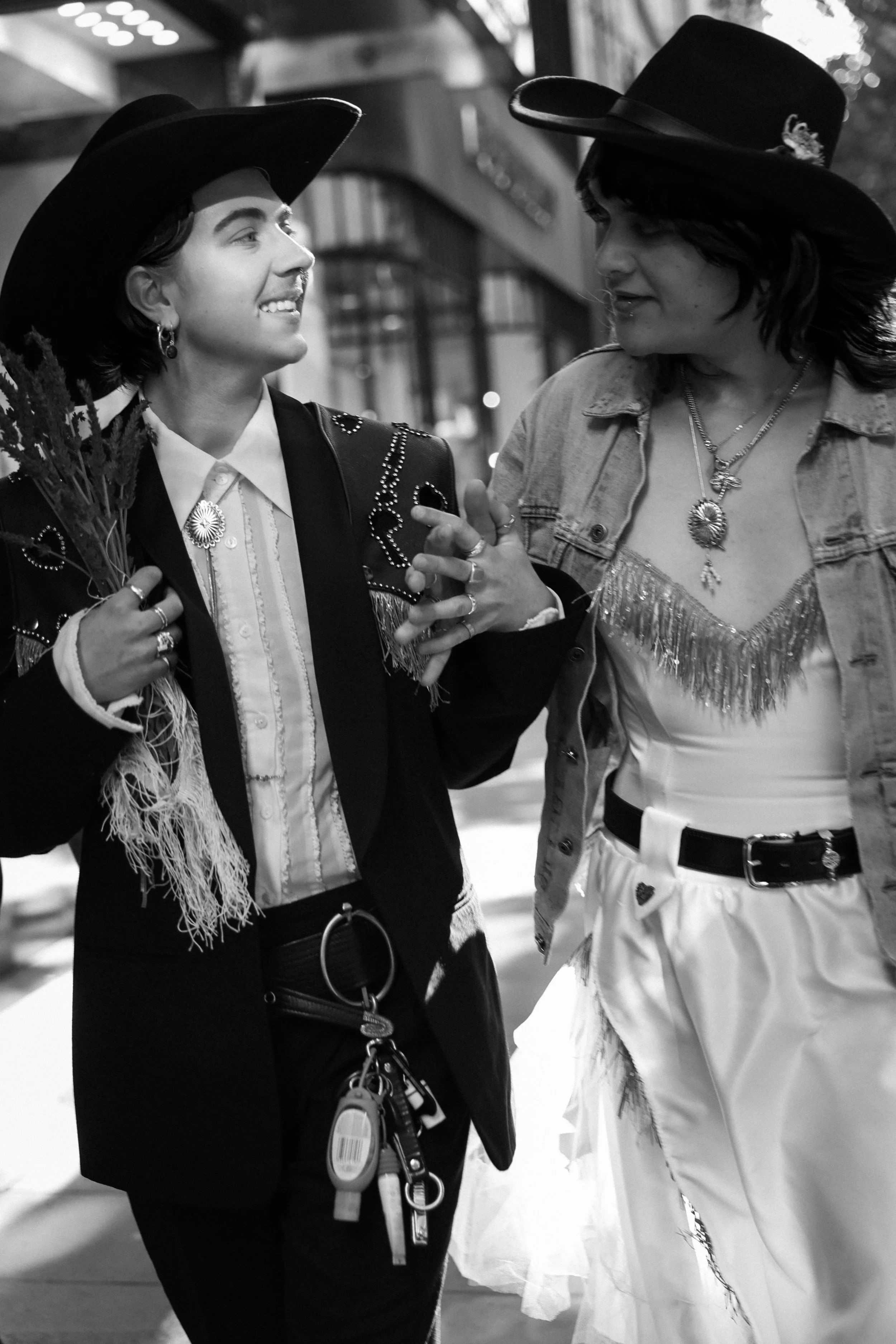 Two women wearing western-style outfits and hats, engaging in conversation on a city street at night.