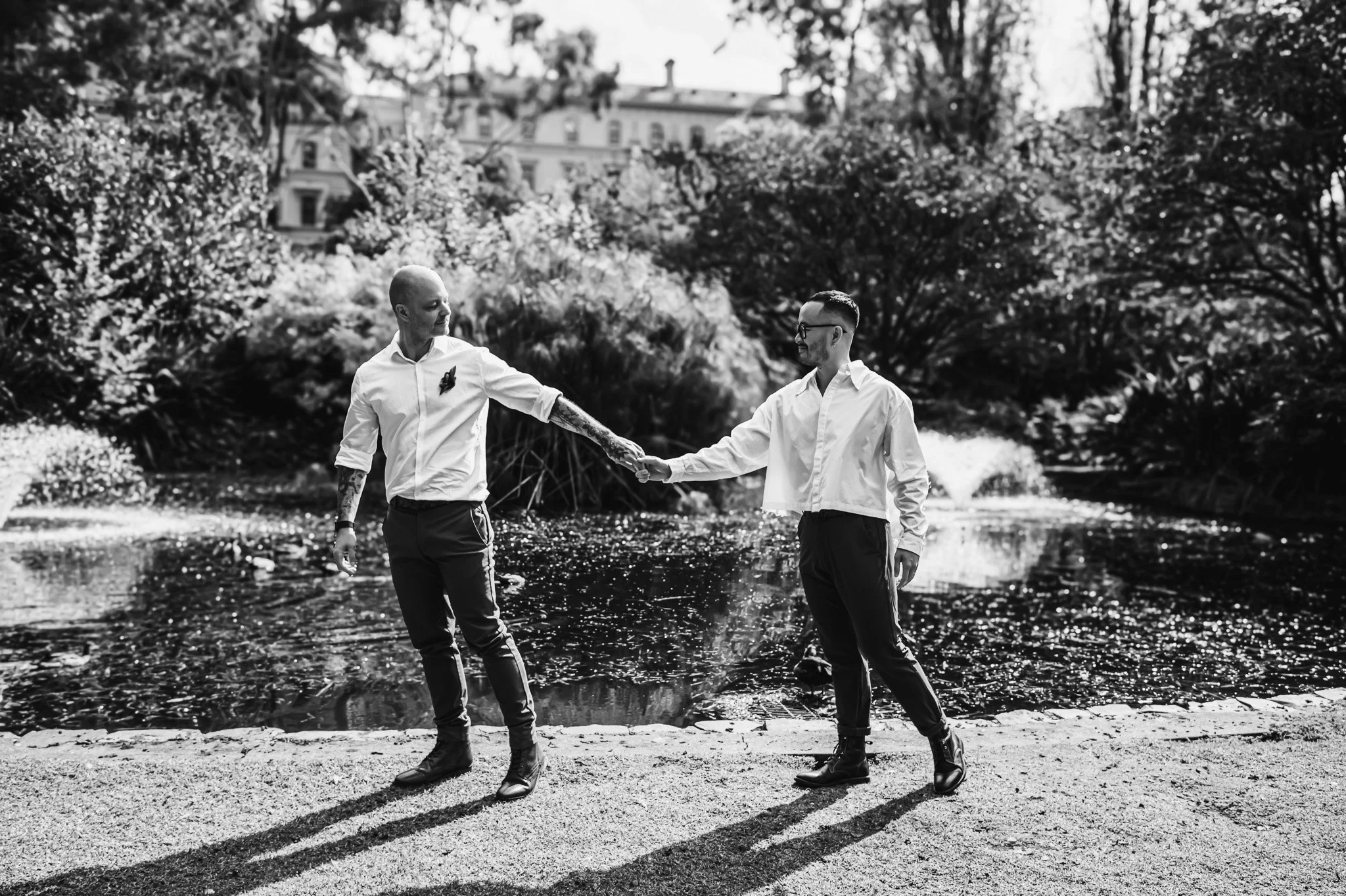 Two men holding hands and walking near a pond in a park, with trees and buildings in the background, black-and-white photo.
