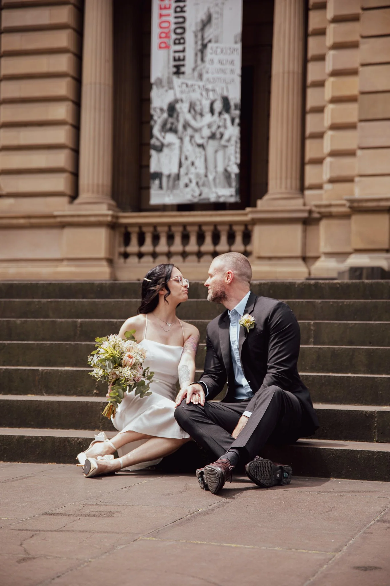 A newlywed couple sitting on the steps of a historic building, sharing a romantic moment, with a large protest banner in the background.