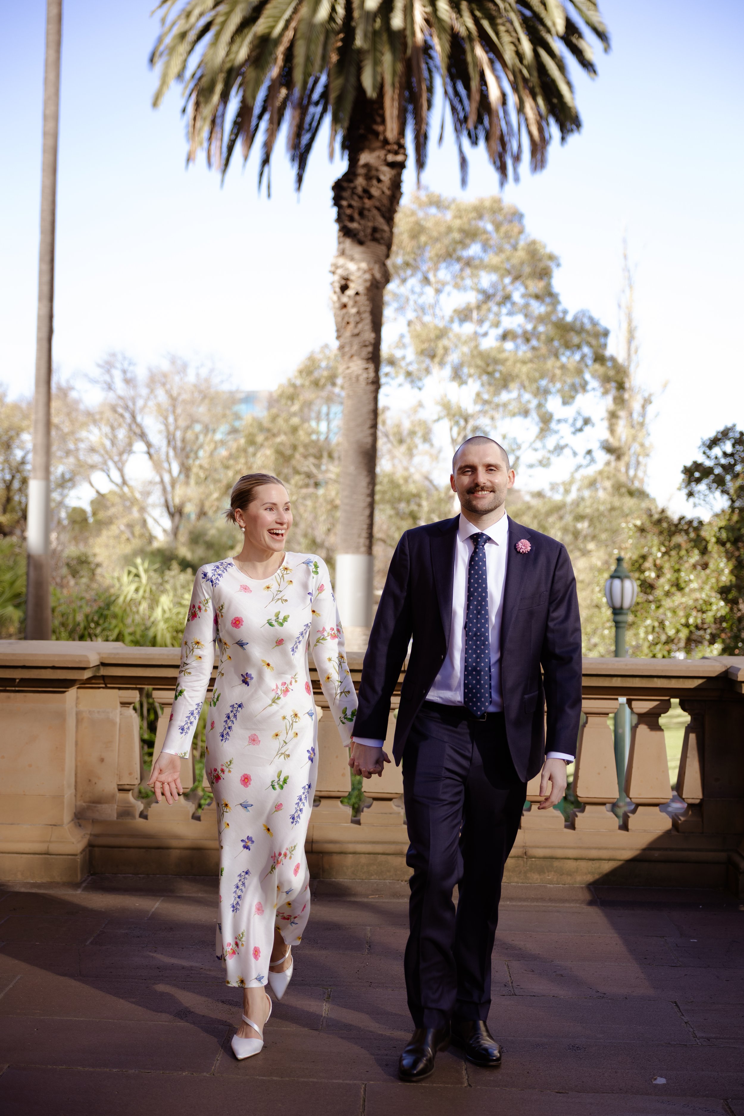 A happily married couple walking hand in hand outdoors, with a tall palm tree and blue sky in the background. The woman wears a white floral dress, and the man wears a dark suit with a tie.