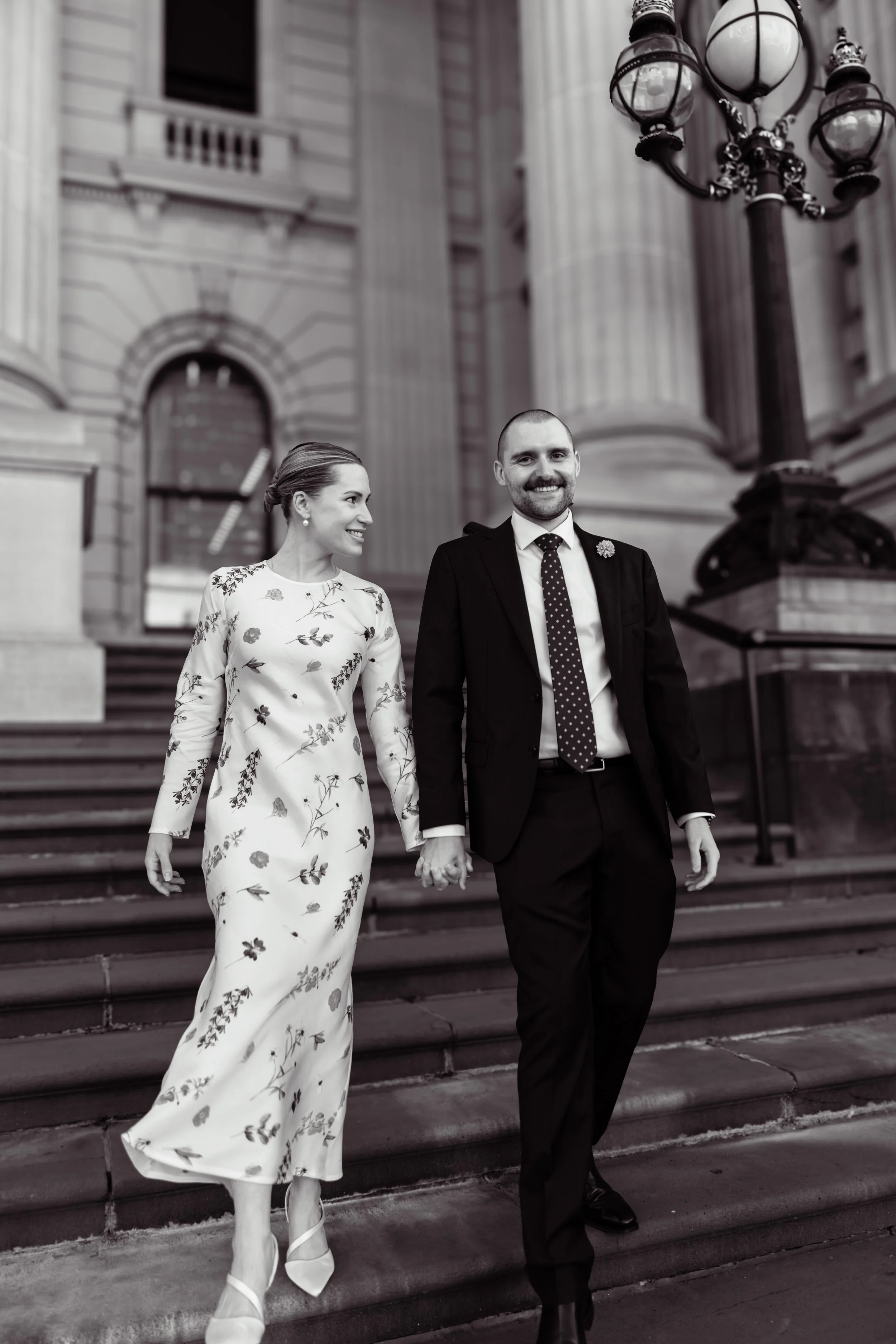 A smiling woman in a floral dress holding hands with a smiling man in a suit walking down city stairs, with a large ornate streetlamp and historic building in the background.