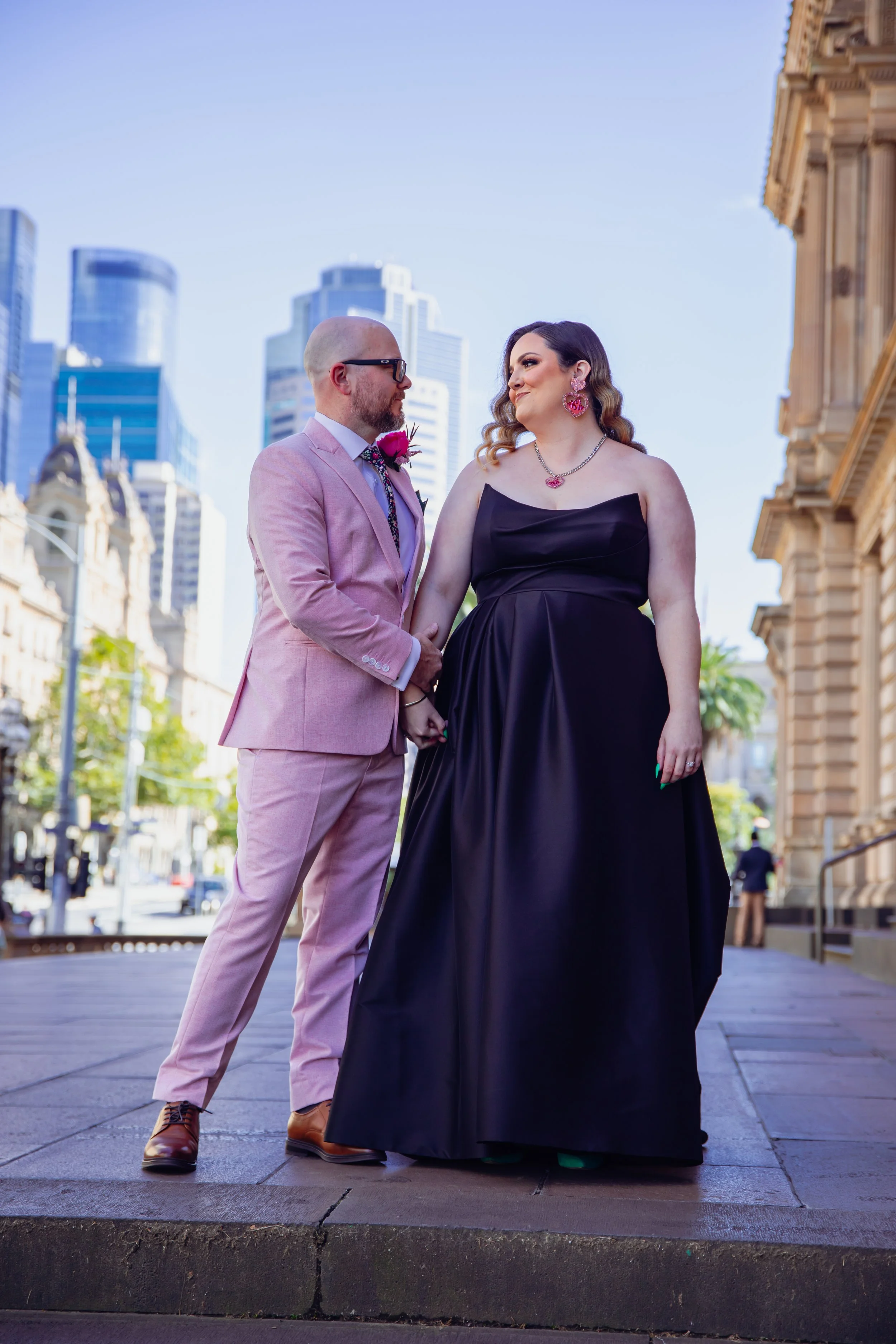 A dressed-up couple holding hands and gazing at each other on a city street with tall buildings in the background.