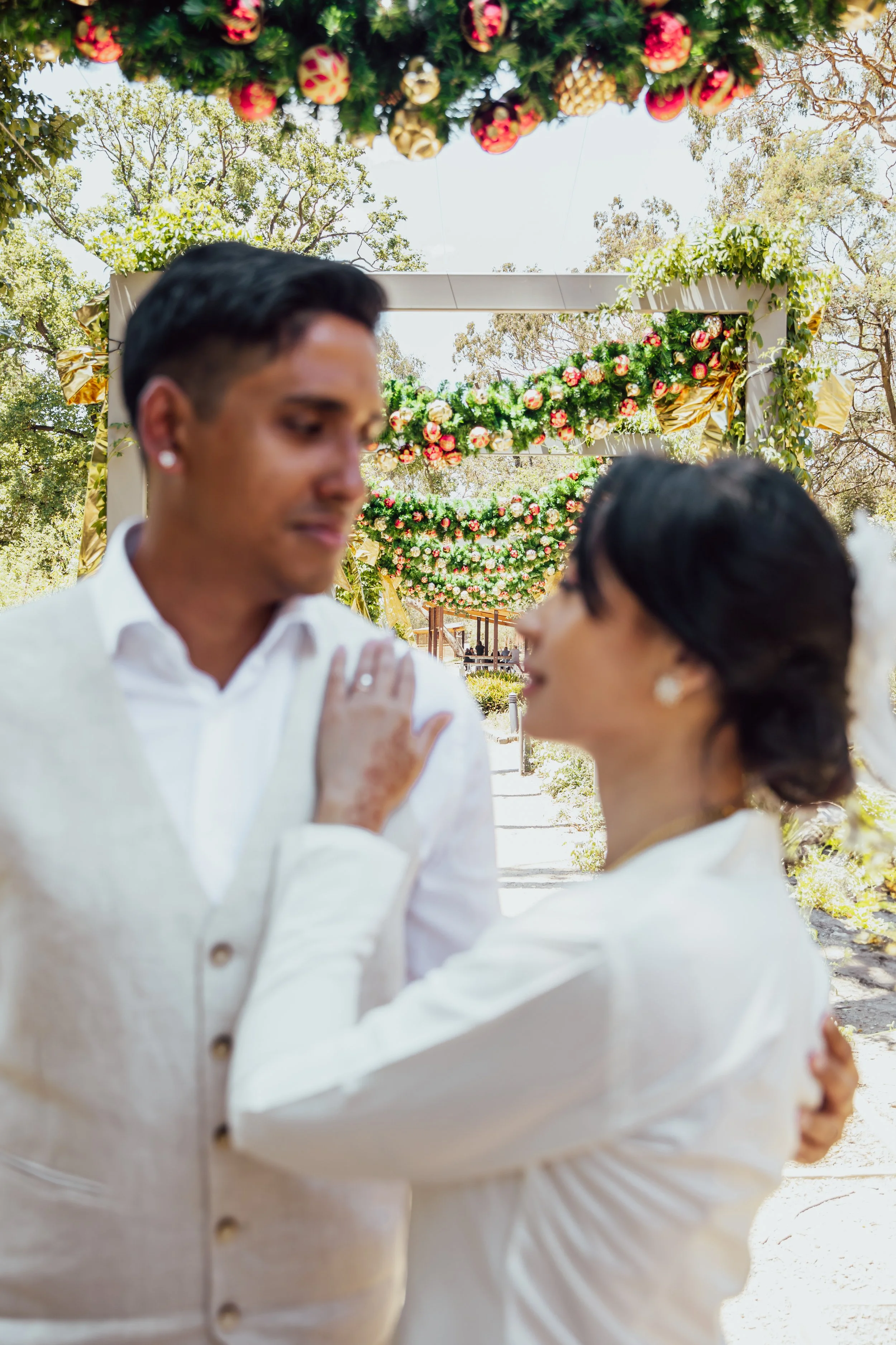A couple dancing at their wedding outdoors under floral archways with hanging flowers, surrounded by trees on a sunny day.