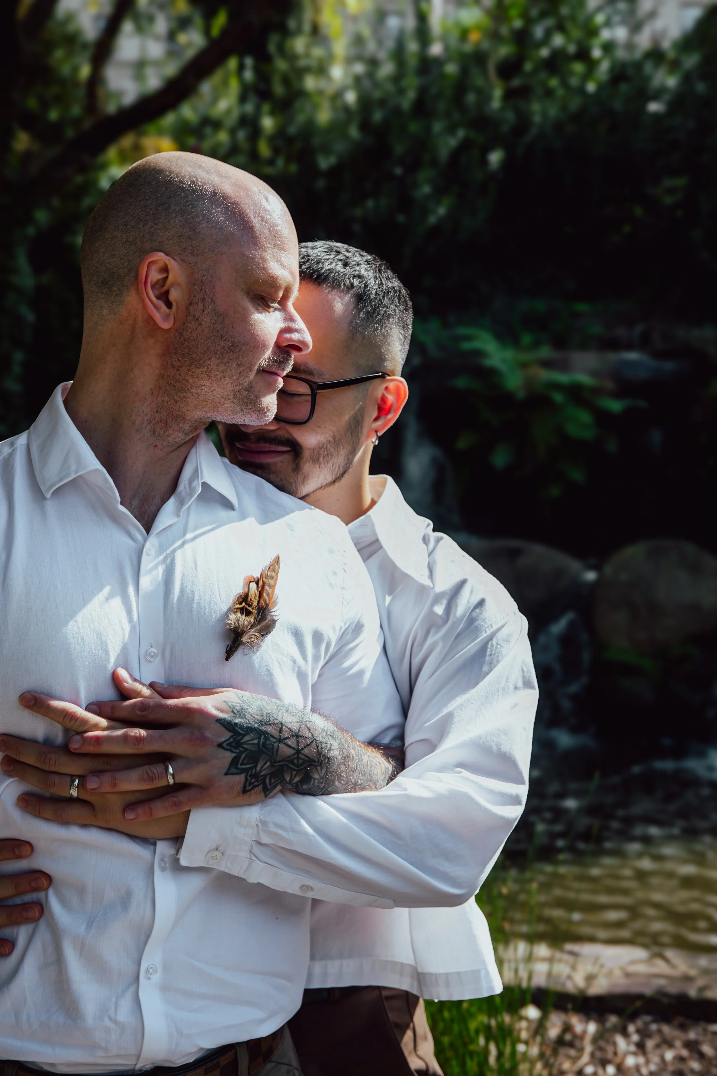 Two men embracing outdoors, one with a tattooed arm and a feather pinned to his shirt, the other with glasses, in a natural setting near water.