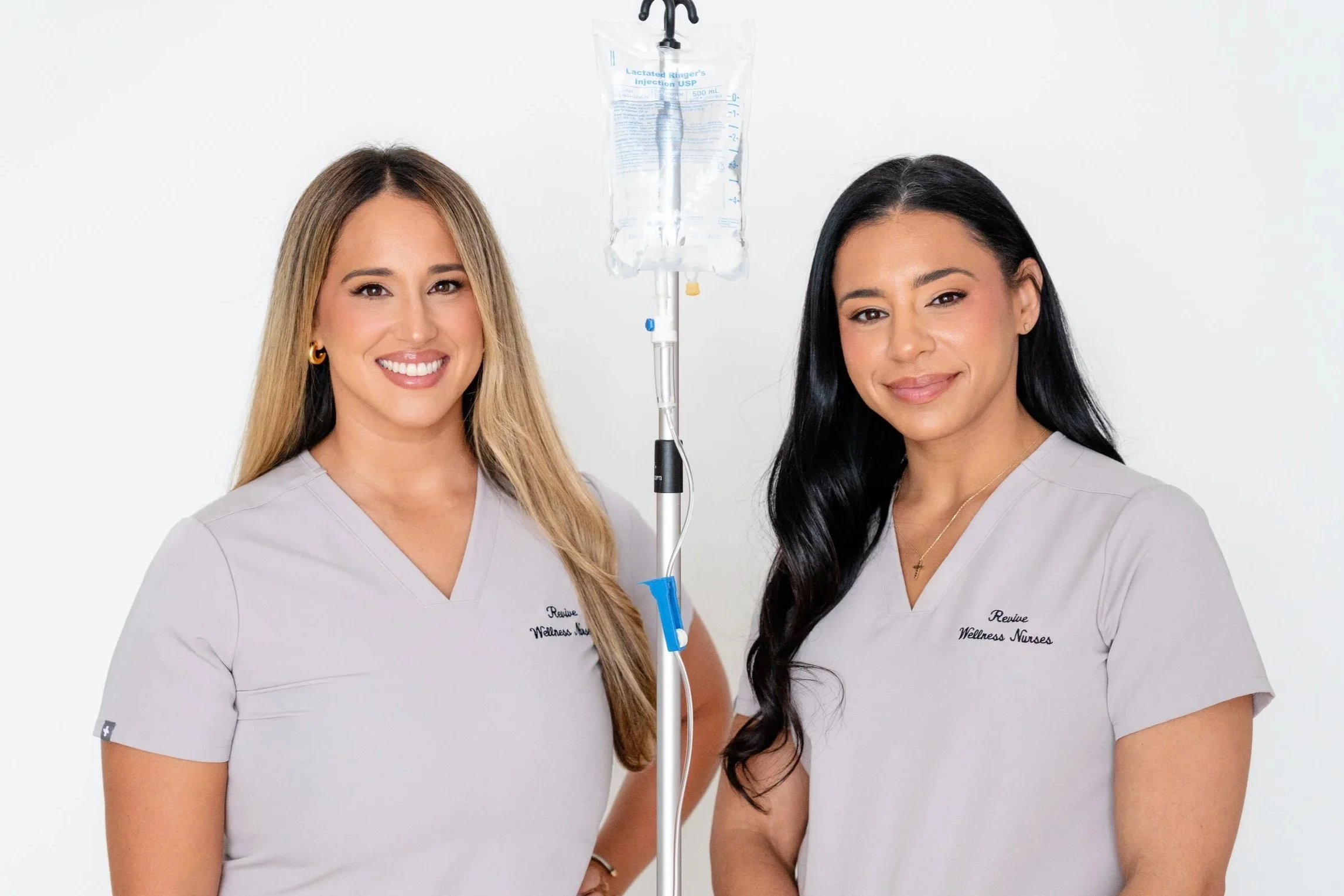 Two women in healthcare uniforms smiling, standing next to an IV drip