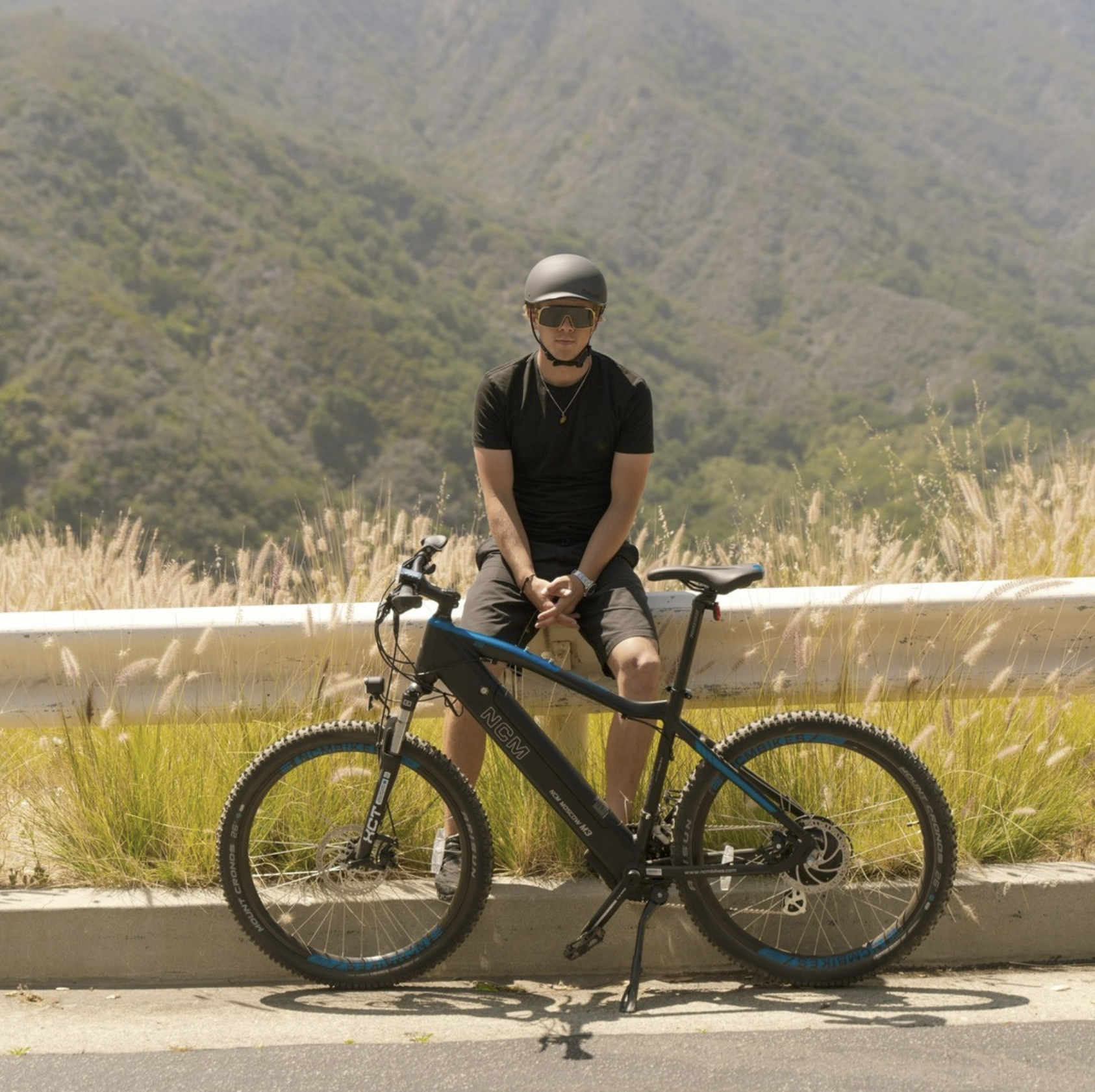 A person wearing a helmet and sunglasses sitting on a mountain bike on the side of a road with a mountainous landscape in the background.