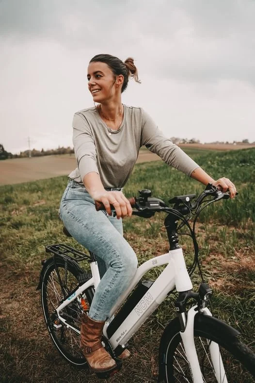 A woman in casual clothing riding an electric bike outdoors in a rural landscape with fields and cloudy sky.