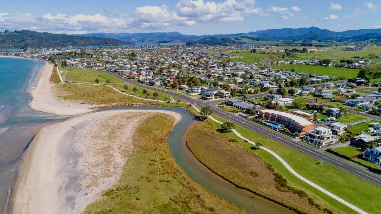 Aerial view of a coastal town with a beach, ocean, a small river, residential houses, and green hills in the background on a partly cloudy day.