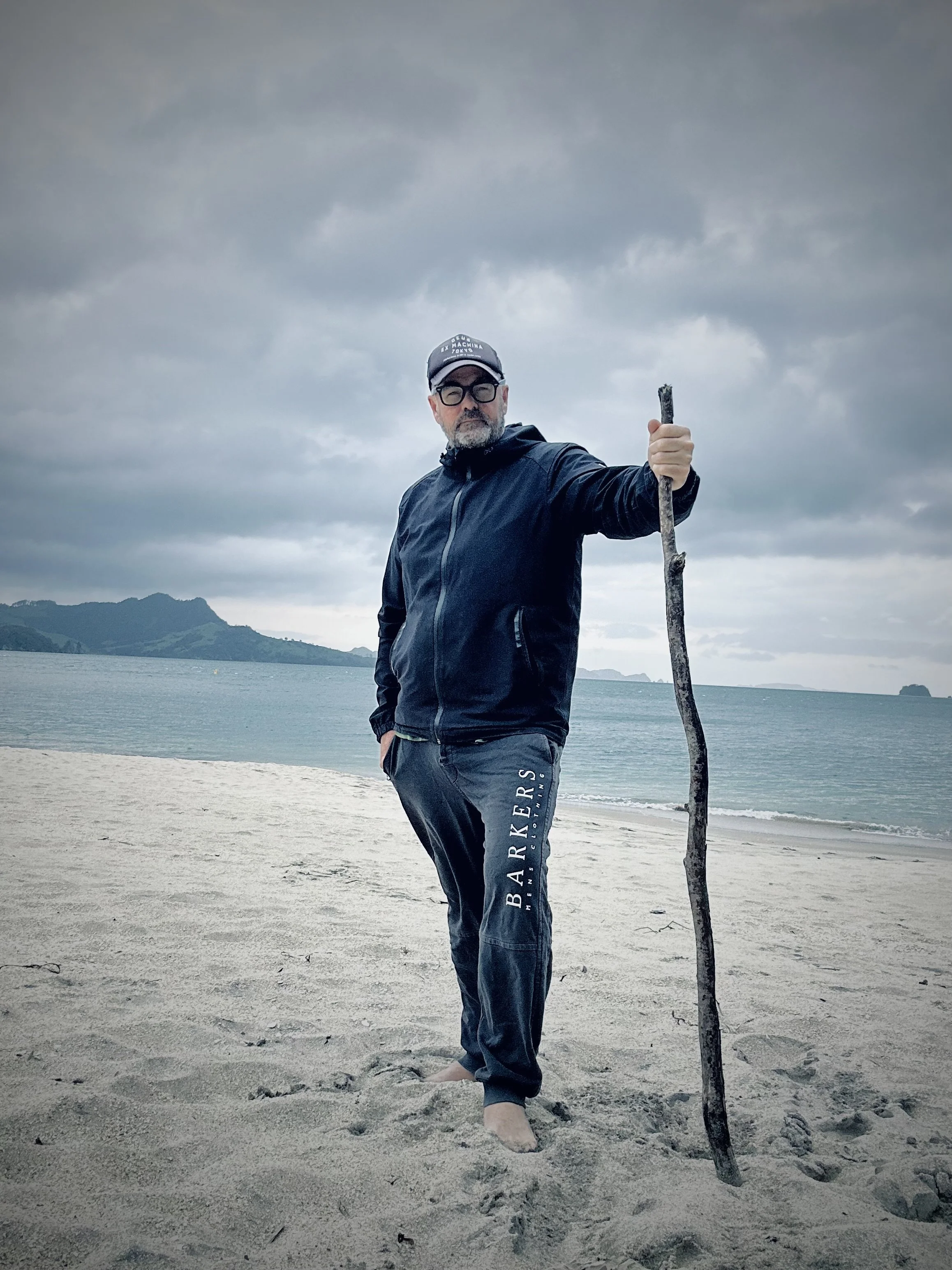 A man standing barefoot on a sandy beach holding a long walking stick, wearing a dark jacket, matching pants with "BARKERS" written on them, a cap, and glasses, with a cloudy sky and mountains in the background.
