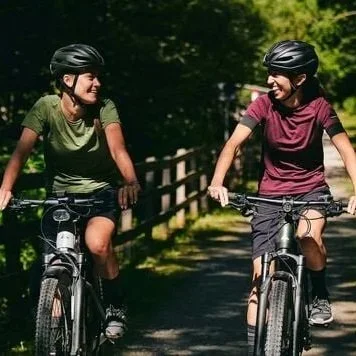 Two women biking on a trail surrounded by trees, wearing helmets and casual clothing, smiling at each other.