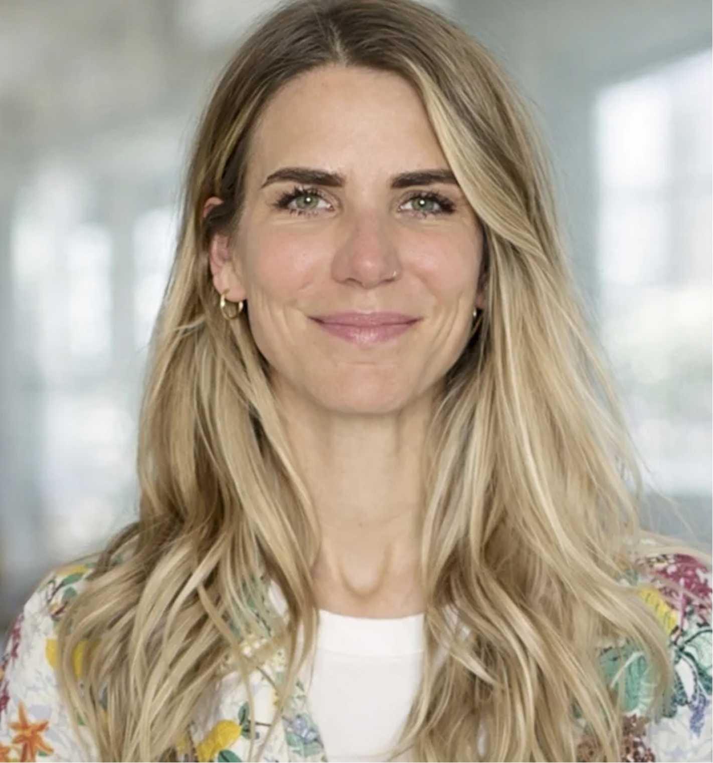 A woman with long wavy blonde hair, wearing hoop earrings and a colorful patterned top, smiling at the camera in a well-lit indoor setting.