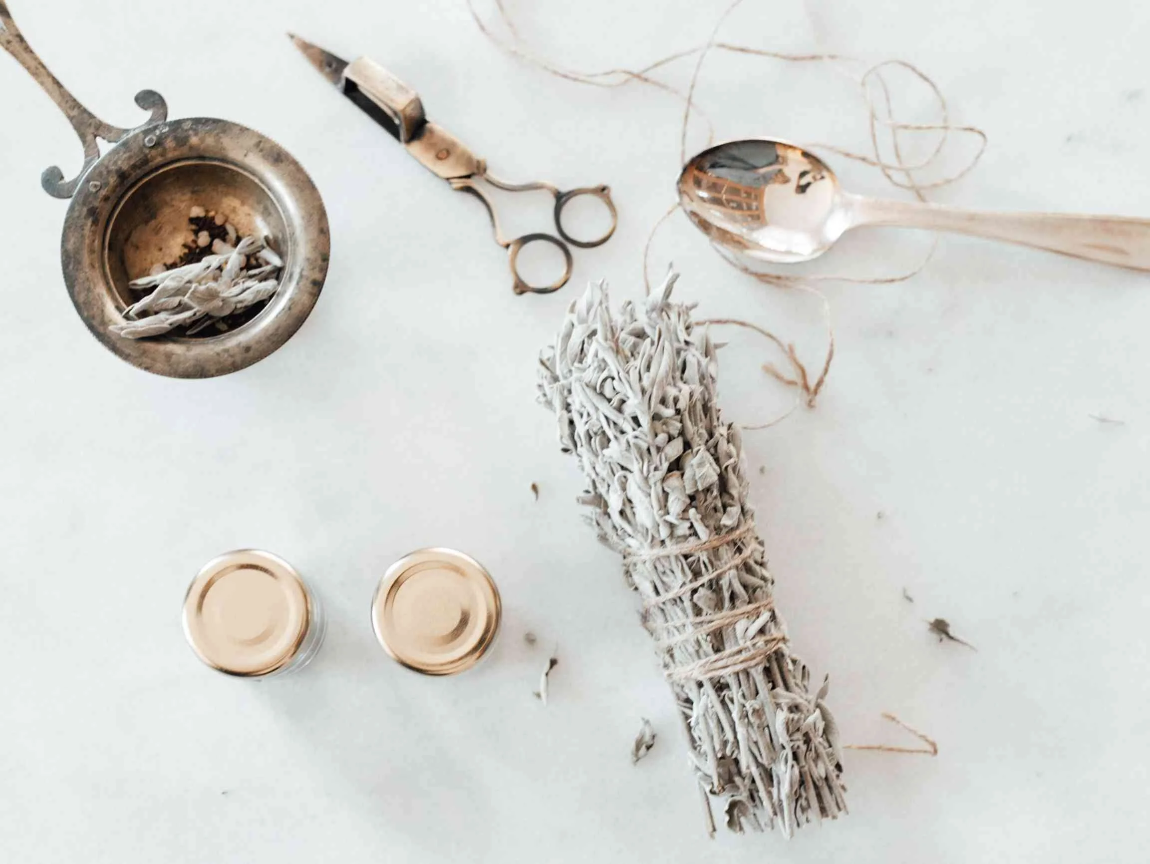 A collection of objects including a bundle of sage, a silver spoon, a pair of cuticle scissors, three small candles, and a brass container filled with dried herbs, all arranged on a white surface.
