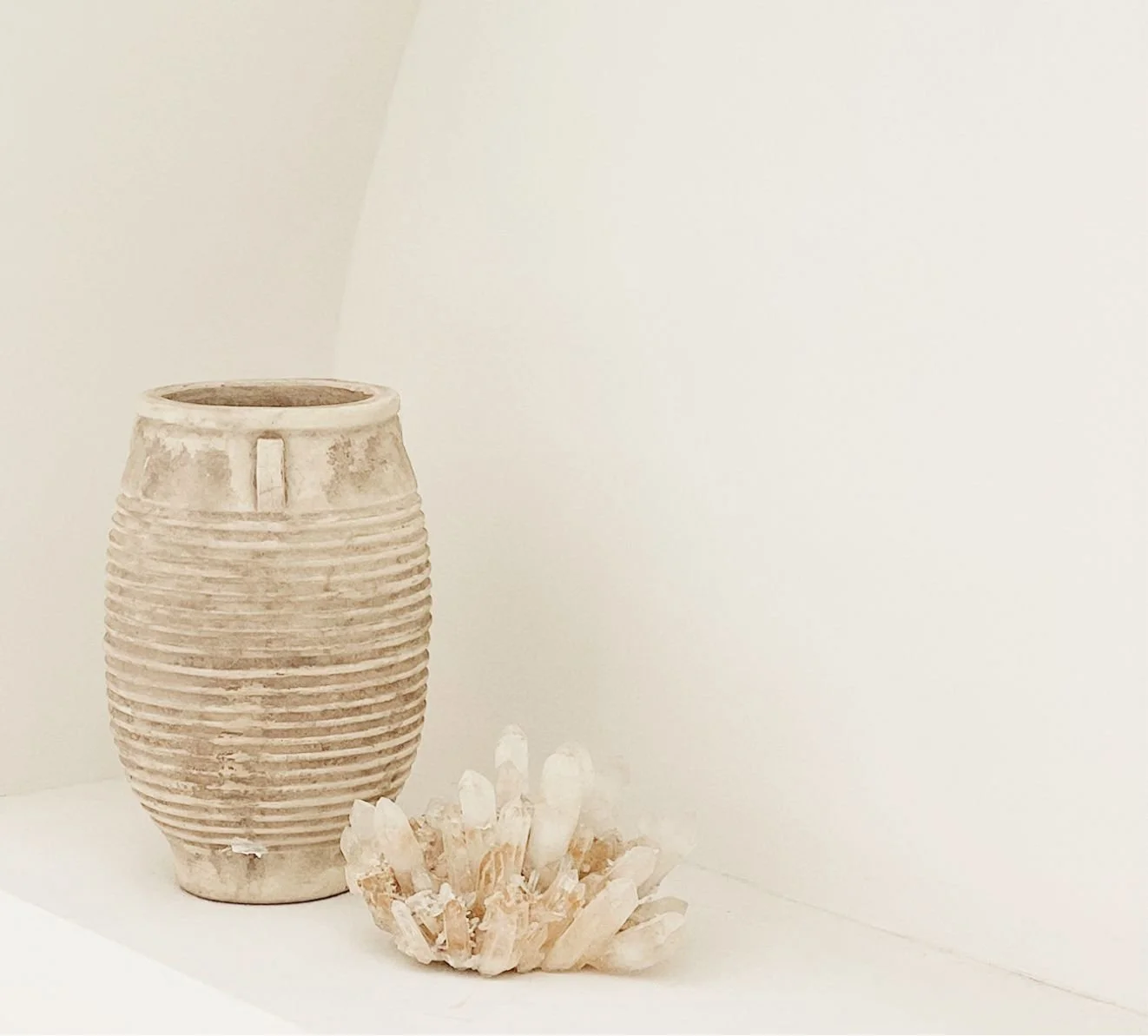 Ceramic vase with horizontal ridges next to a cluster of quartz crystals on a white surface and wall background.