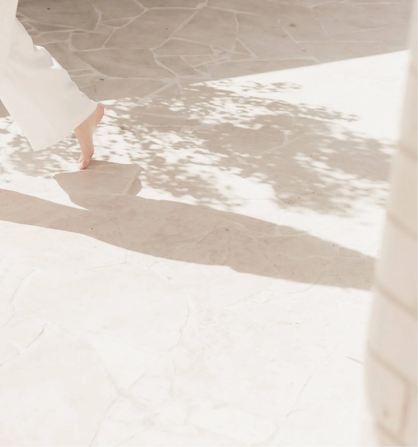 A person wearing a white shirt walking on a tiled floor, with shadows of nearby foliage cast on the ground.