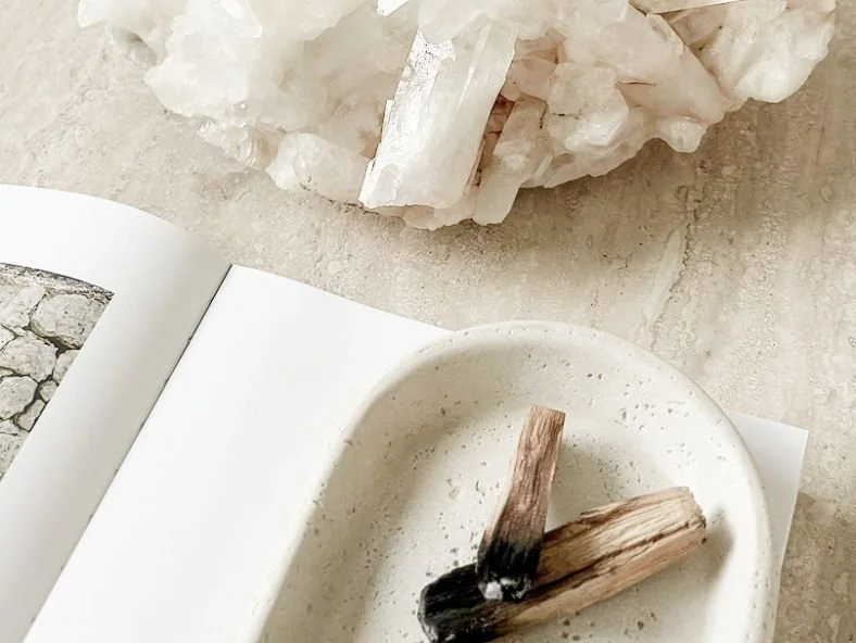 A beige marble surface with a white open book, a round white plate containing burnt wood sticks, and a large cluster of white crystals or minerals.