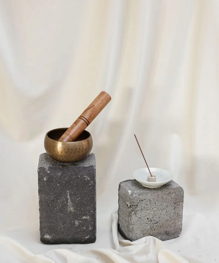 Two concrete blocks with Asian musical instruments on top, a singing bowl with a wooden striker and an incense holder with a stick of incense.