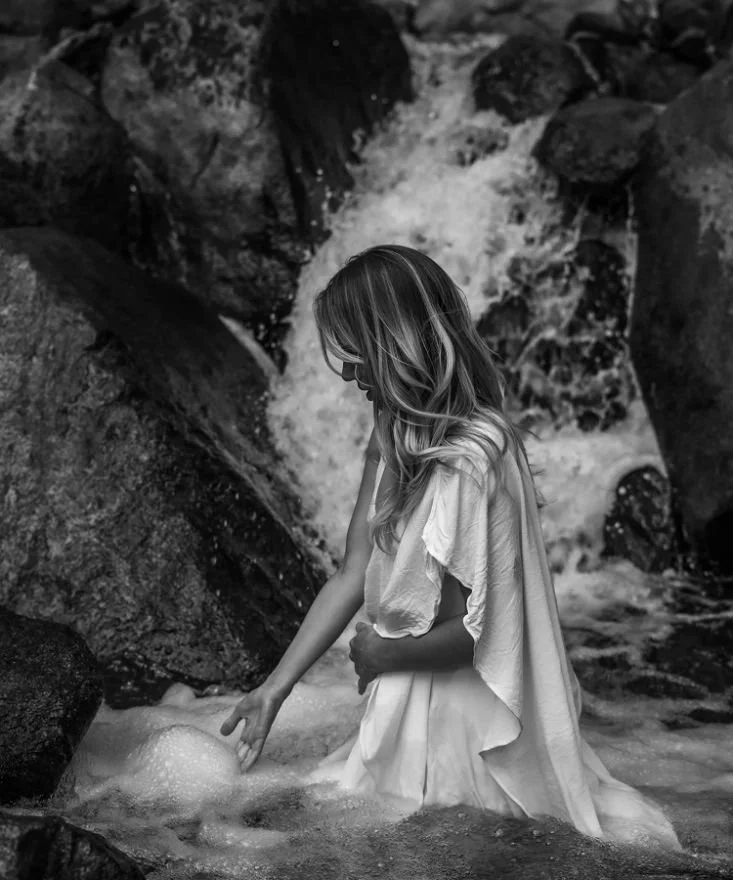 A woman with long hair in a white dress kneels in a rocky stream by a small waterfall, touching the water.