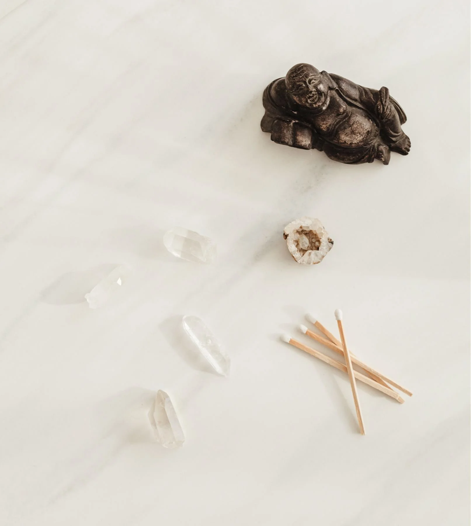 A small black figurine of a laughing Buddha, a clear quartz crystal, a geode, and a bundle of matchsticks on a white surface.