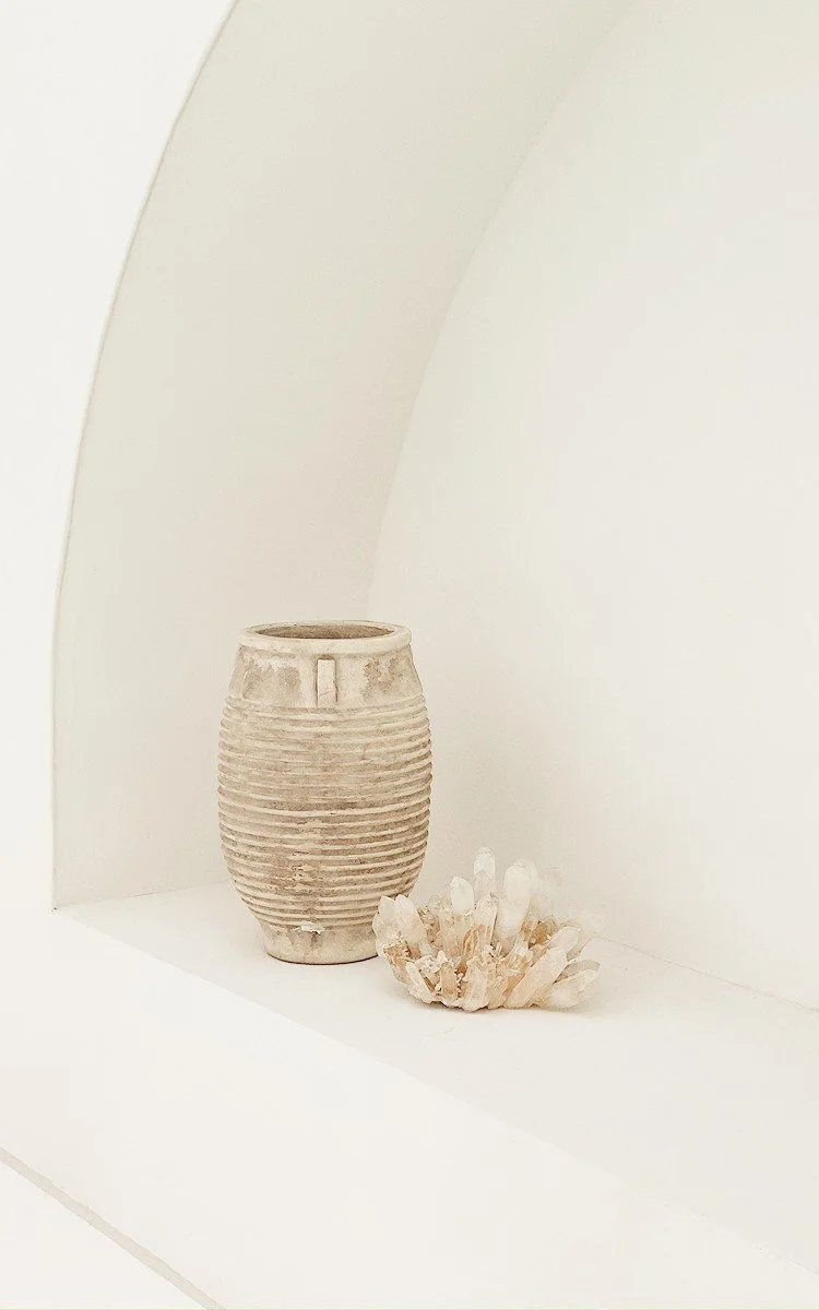 A textured beige ceramic vase and a seashell with white crystals on it placed on a white shelf against a white wall with minimalist architecture.