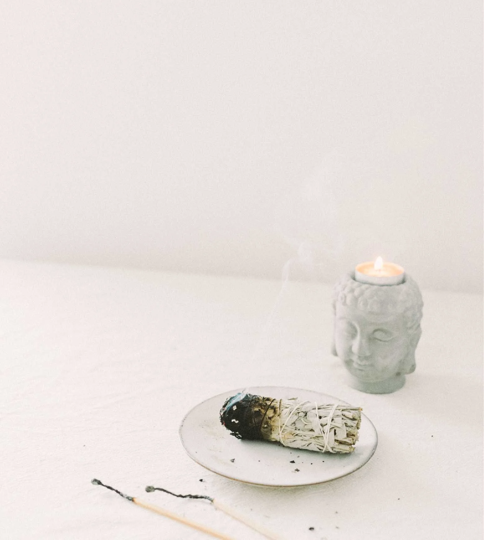 A white tabletop with a burning incense cone with smoke, a small fish-shaped incense holder, and a Buddha head sculpture with a candle on top. A small, unfinished charcoal drawing is on the table.