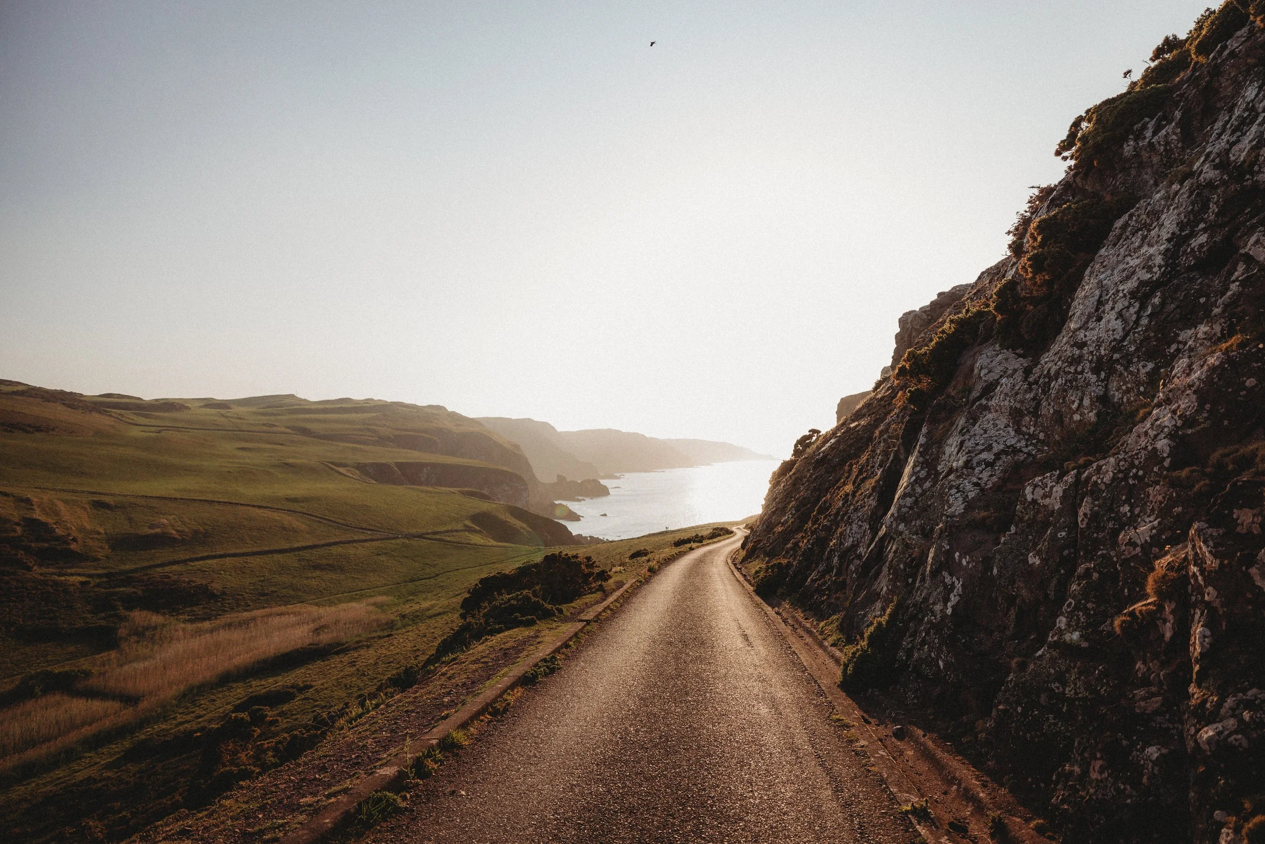 A winding rural road runs through green hills and alongside a rocky cliff, leading towards the ocean in the distance with the sky clear and sunlight illuminating the landscape.