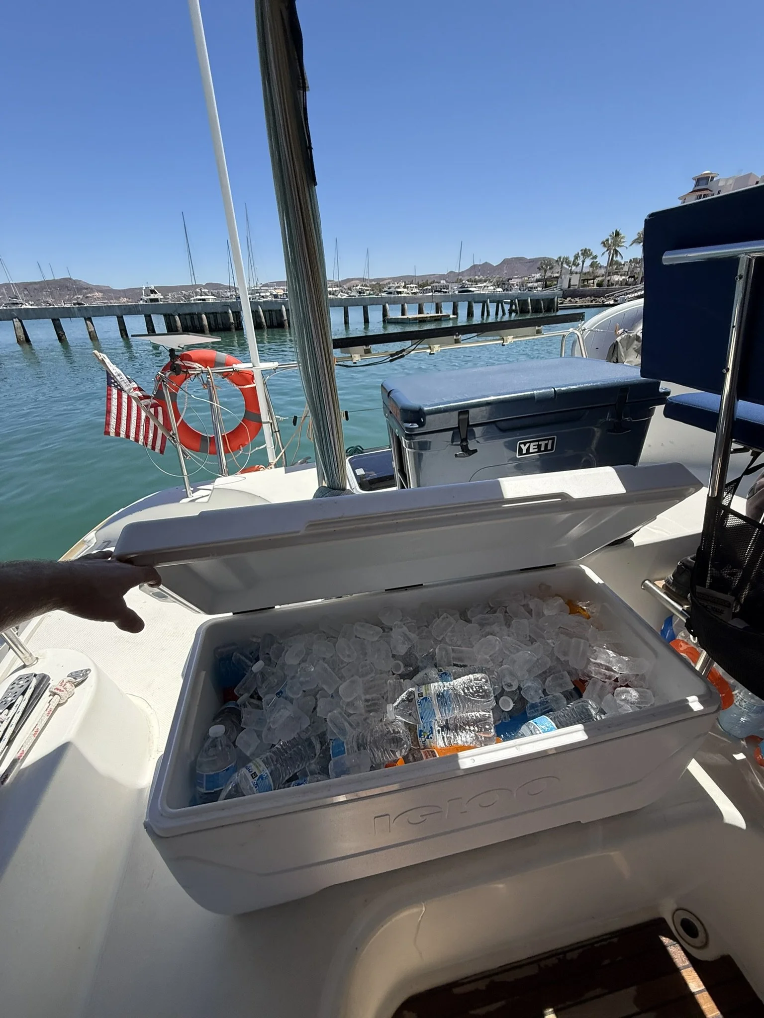 View from a boat showing a cooler filled with ice and water bottles, a dock with boats in the water, and a background of mountains and palm trees under a clear blue sky.
