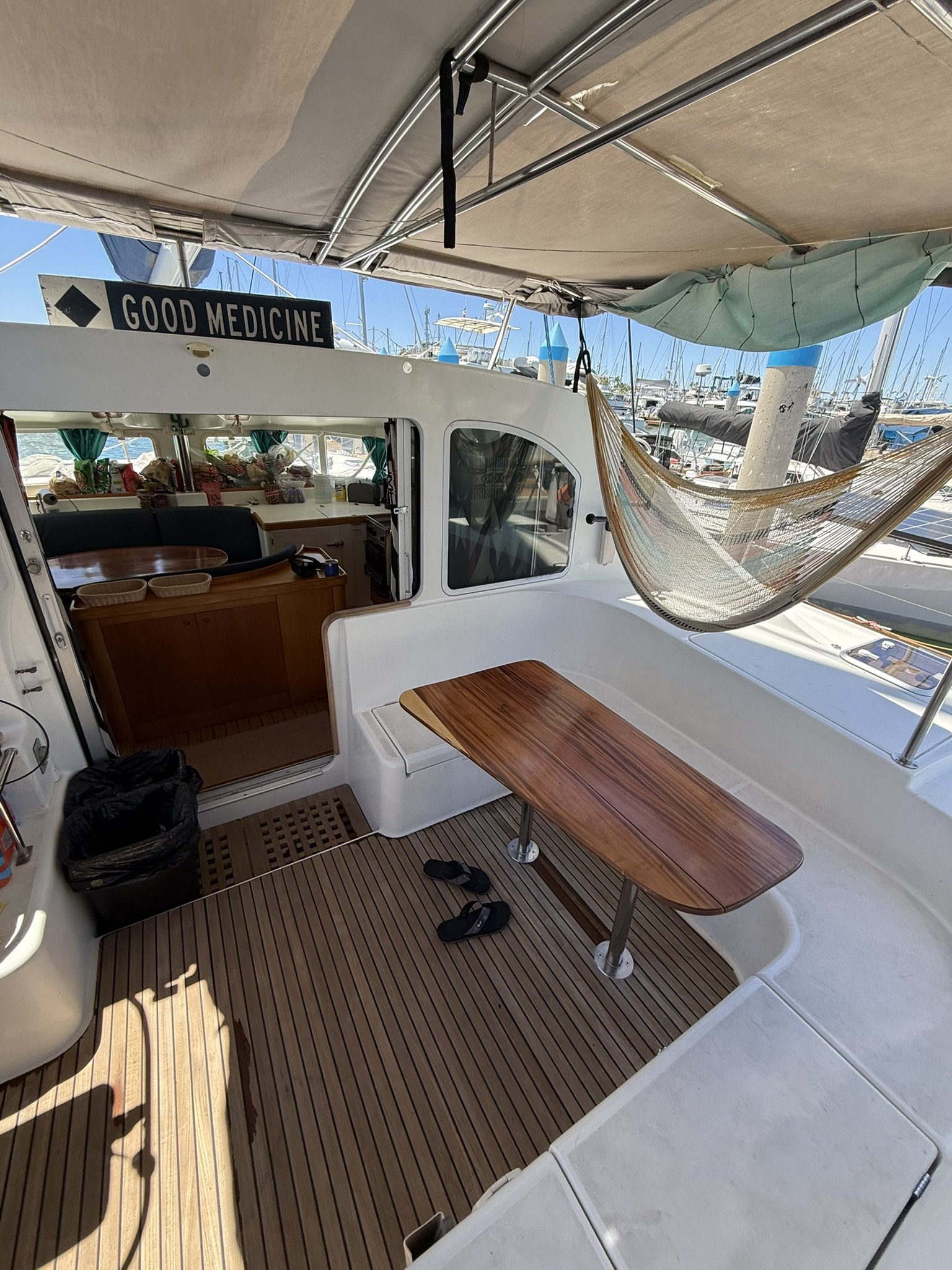 View of a boat with a wooden deck, a table with a pair of sandals underneath, a hammock, and a view of marina with boats and blue sky in the background.